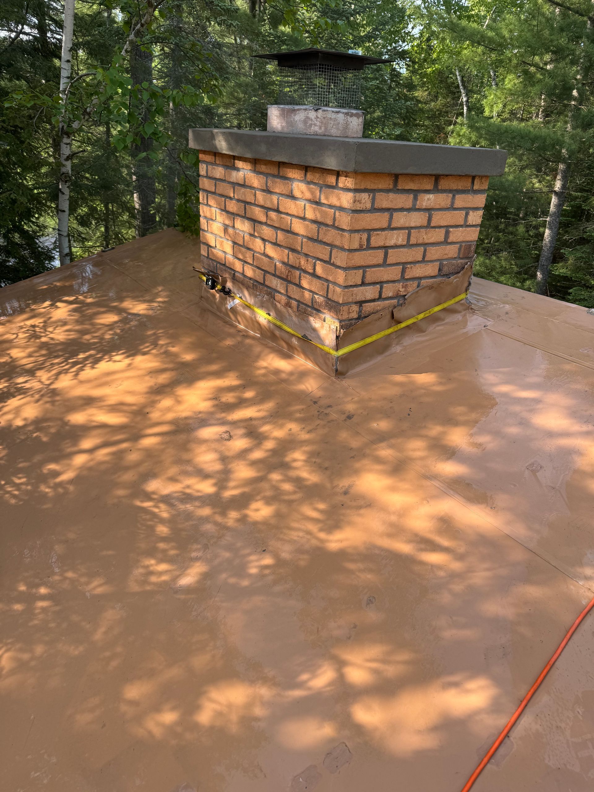 Brick chimney on a brown flat roof surrounded by trees; the roof appears wet.