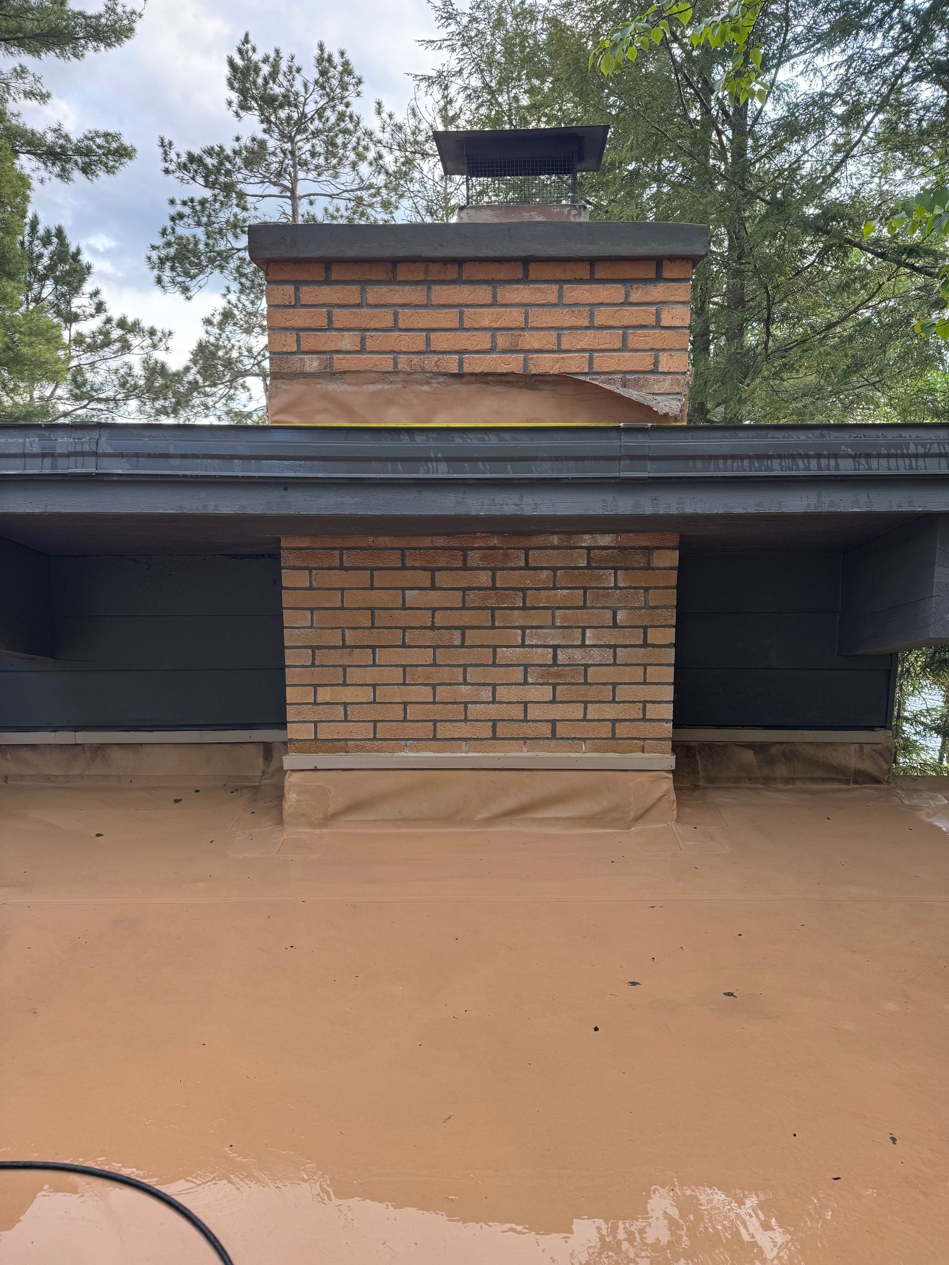 Brick chimney on a flat roof, surrounded by tan-colored material. The sky is visible.