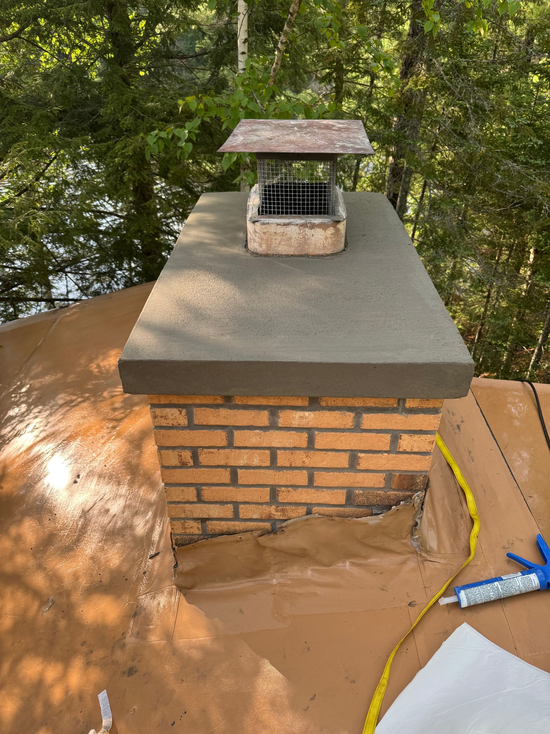 Brick chimney with new concrete cap and metal chimney cap on brown roof.