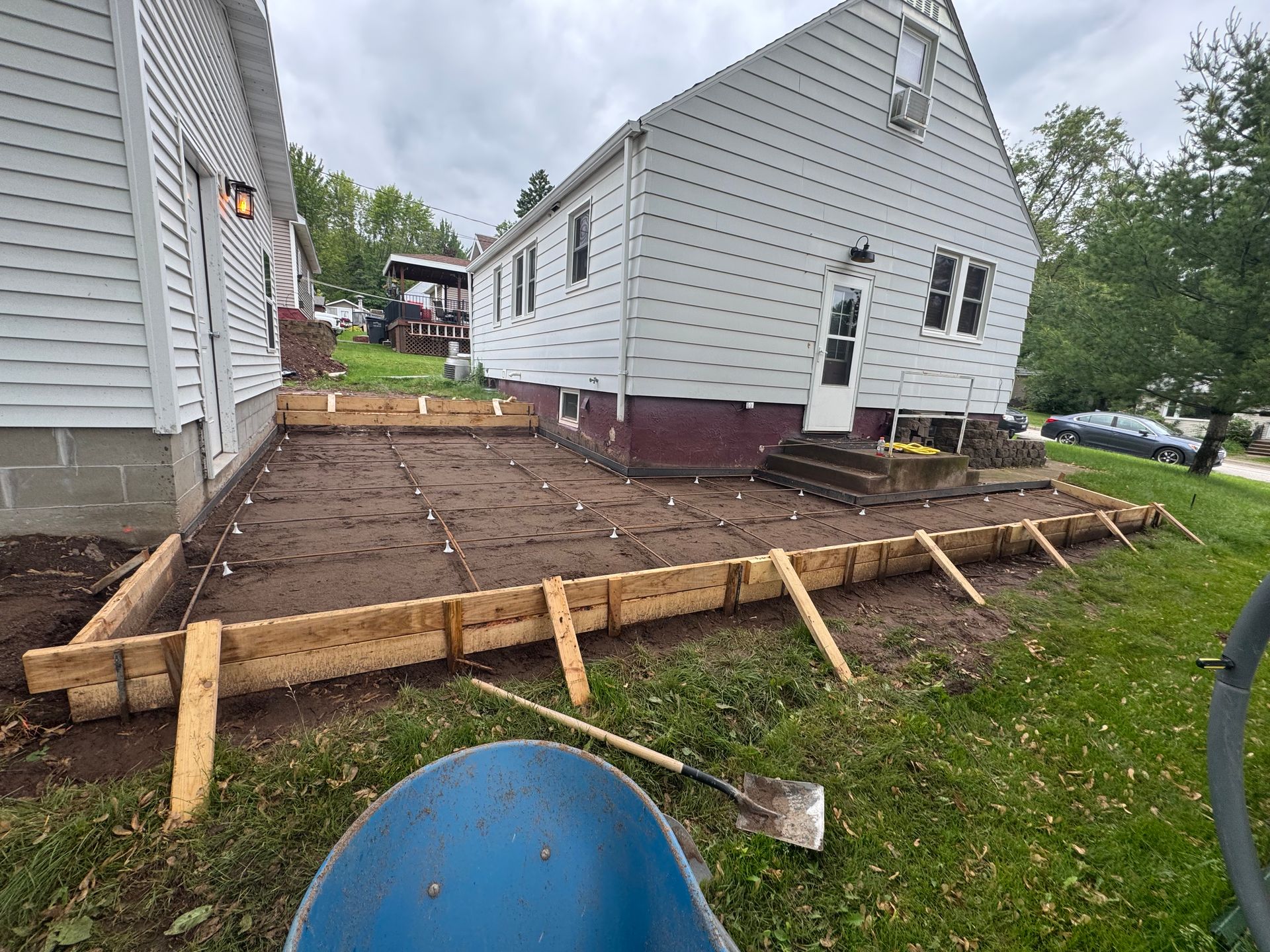 Wooden forms outline a backyard concrete patio project next to a house with white siding and a brown foundation.