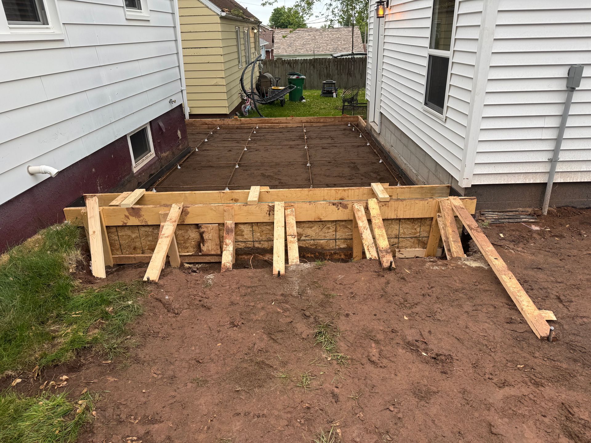 Wooden forms for a concrete patio are being built between two houses; dirt ground.