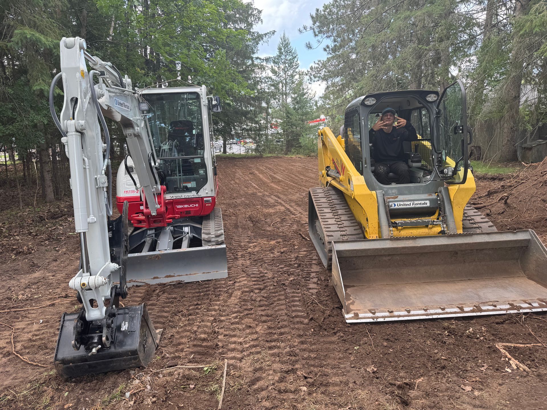 Two construction vehicles on a muddy path: a red and white excavator, and a yellow skid steer with a person inside.