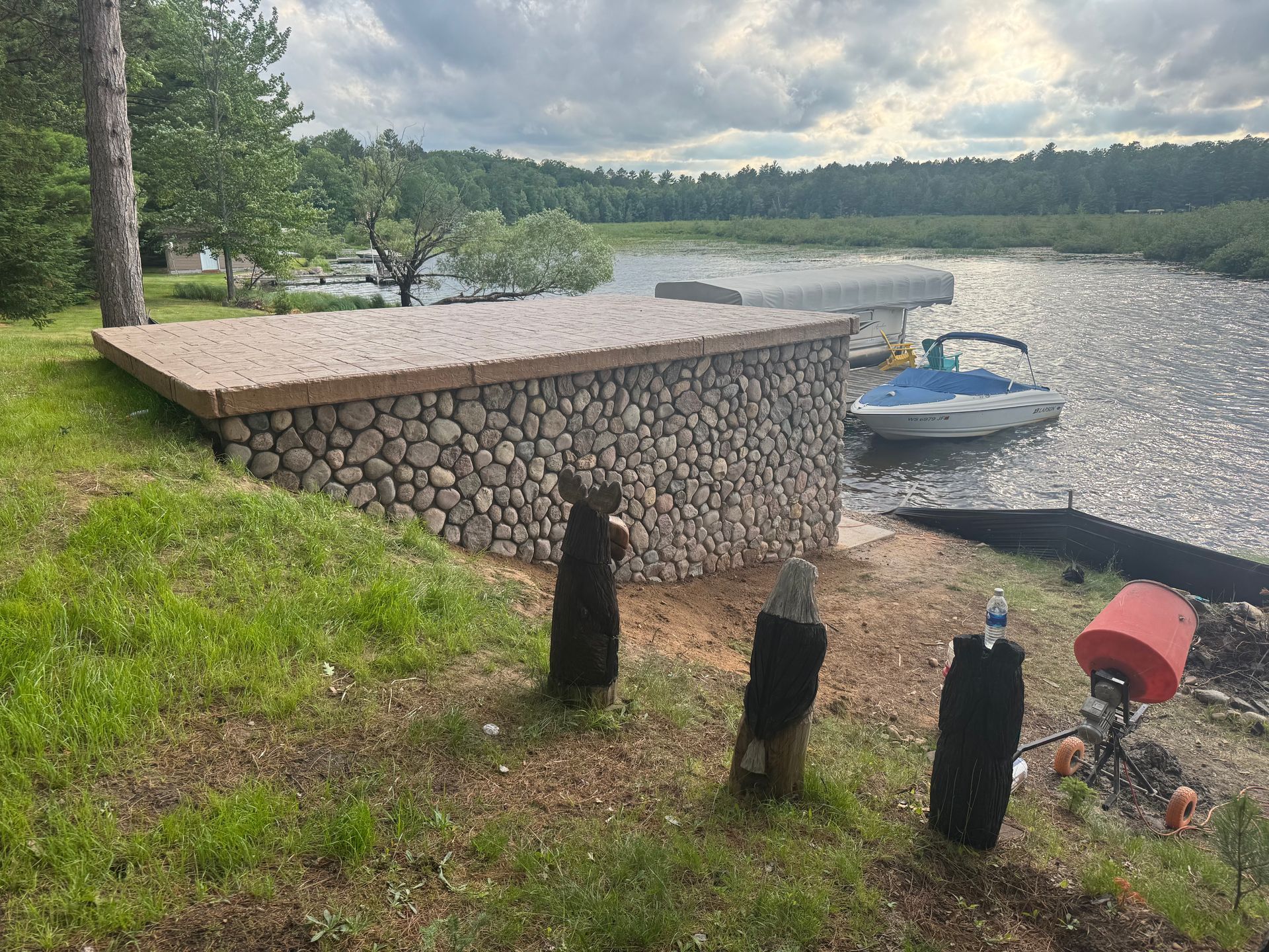 Stone-walled lakeside deck with a boat tied up nearby. Overcast sky, green grass, and water.