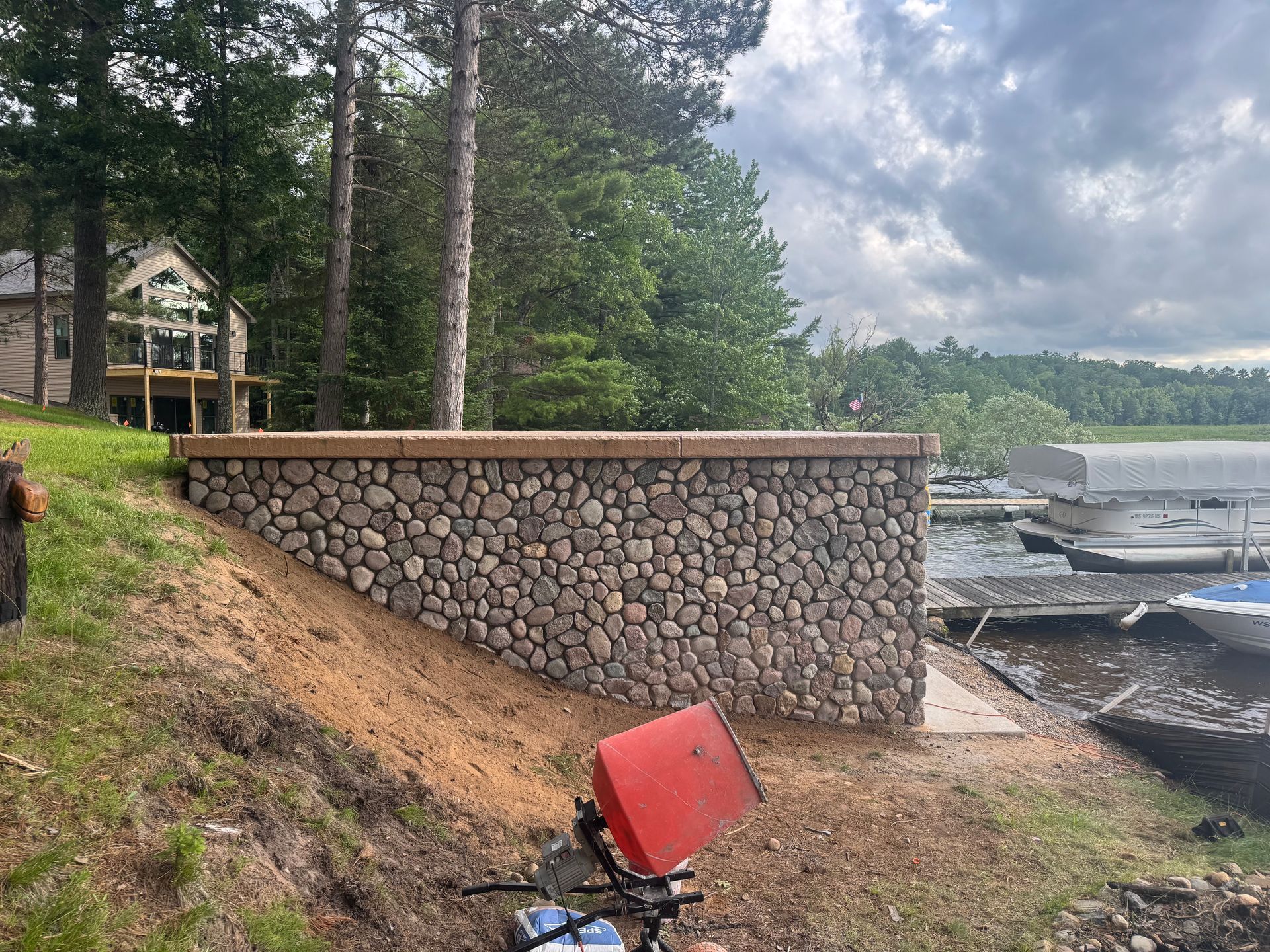 Stone retaining wall near a lake, topped with a tan border. A red wheelbarrow is on the slope in front.