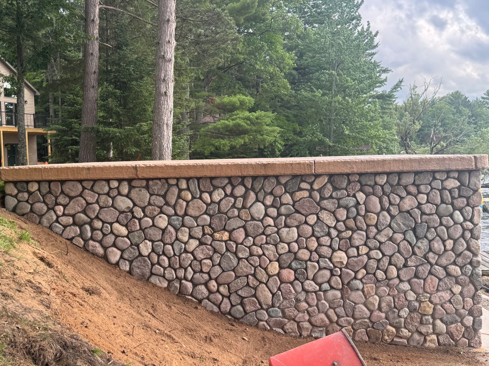 Stone wall with a concrete cap. Bank of dirt slopes downward; trees and a building are in the background.