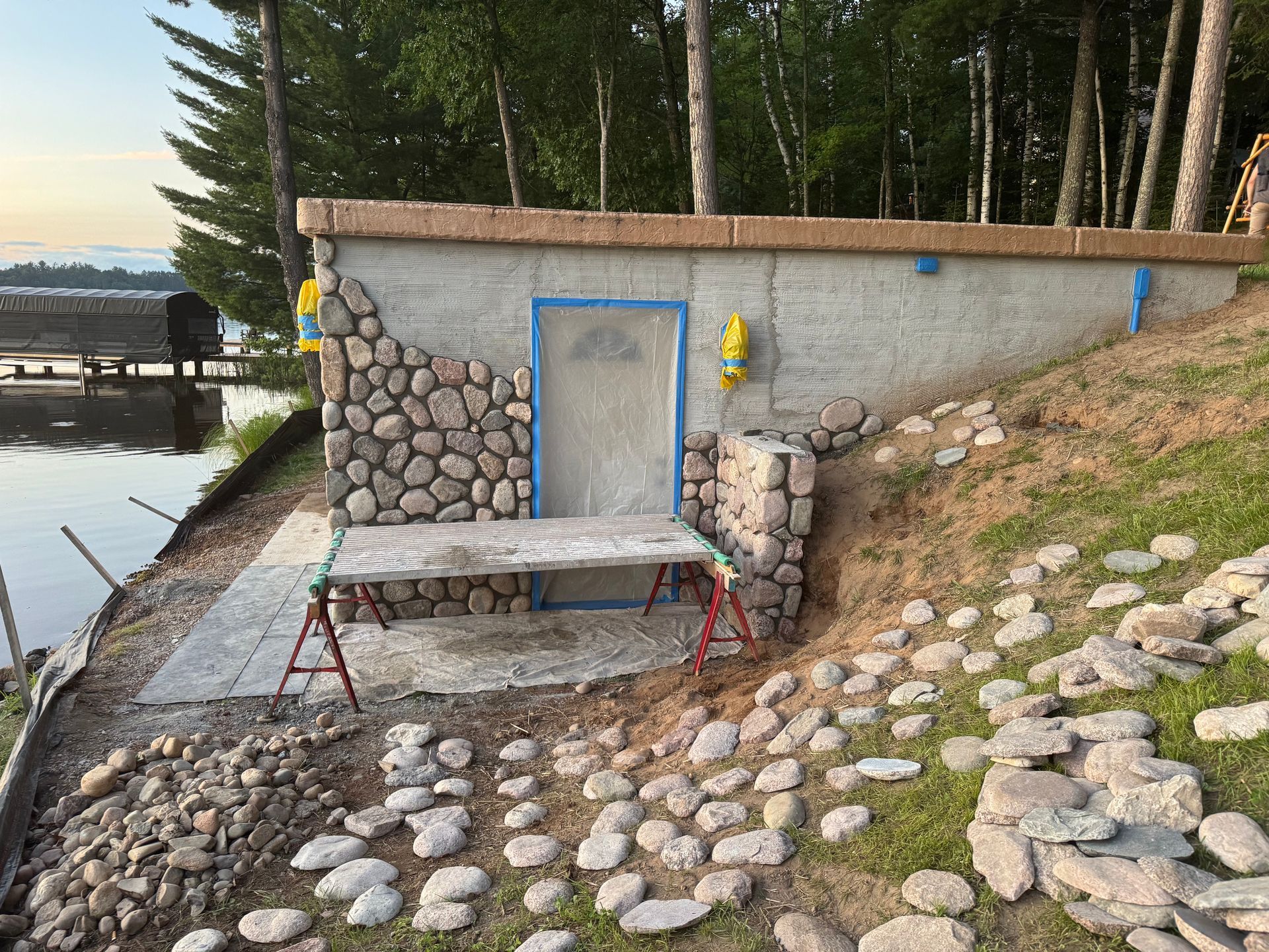 Stone building with stone facade and metal door, next to a lake. A work table and scattered stones are in front.