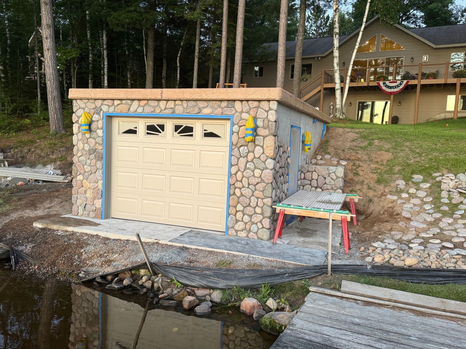 Stone garage by water with tan door, steps, and table; house in the background.