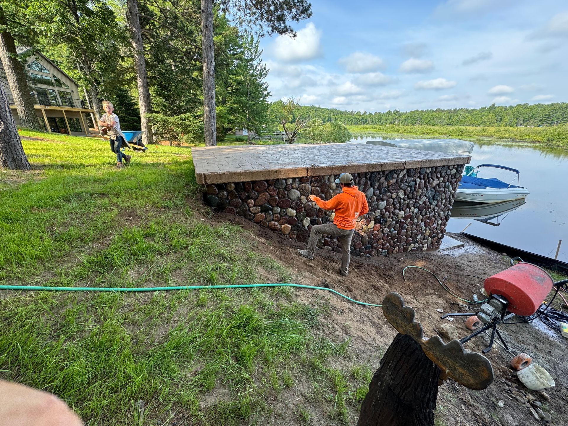 Person working on a stone structure by a lake, another person nearby. Blue sky, greenery.