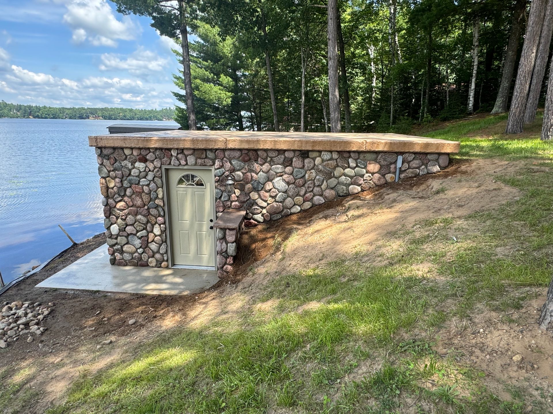 Stone-walled storage building with tan door, concrete base, and brown roof near a lake.