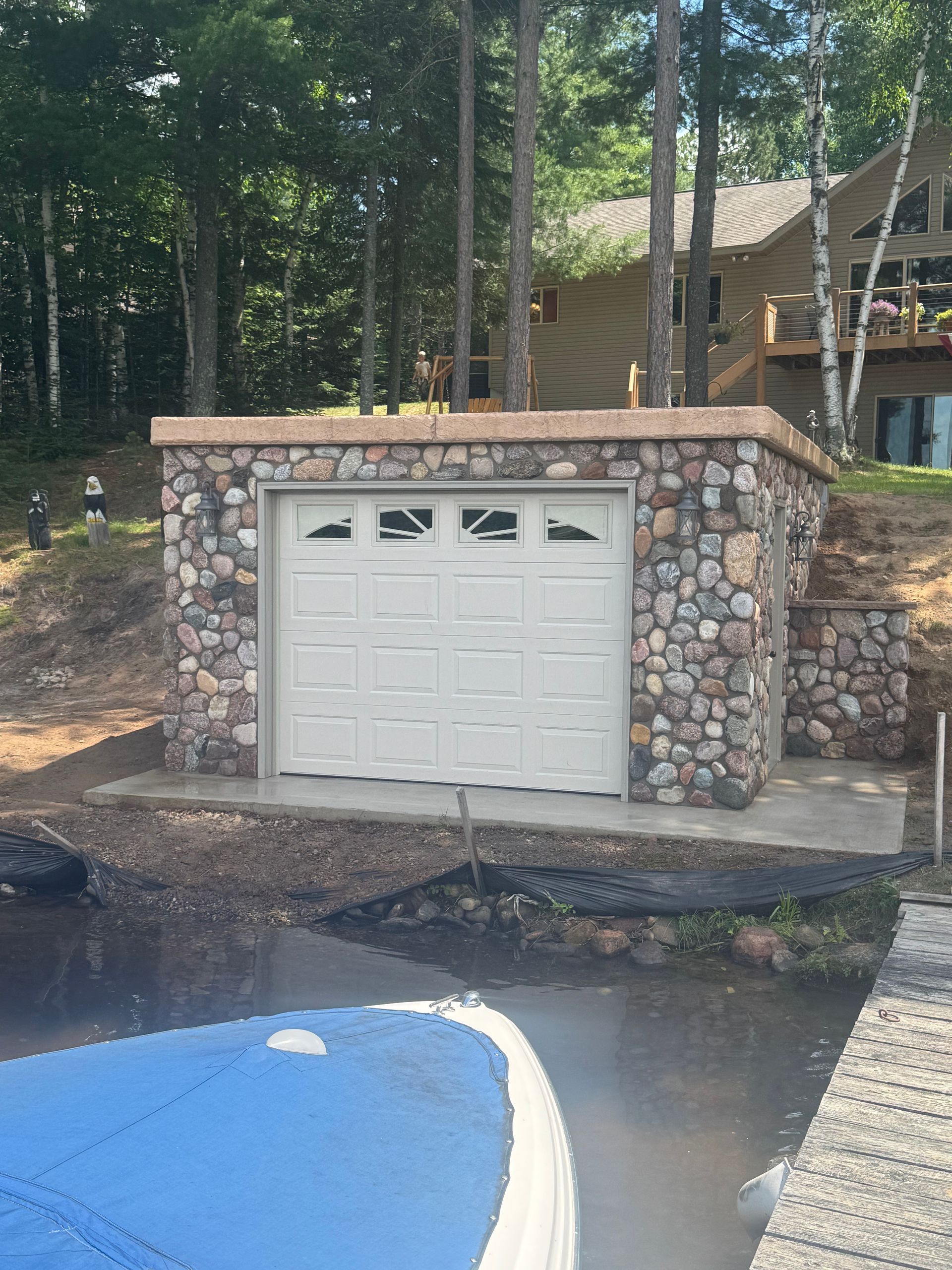 Stone garage on a lake. A blue boat is in the foreground. Trees and a house are in the background.
