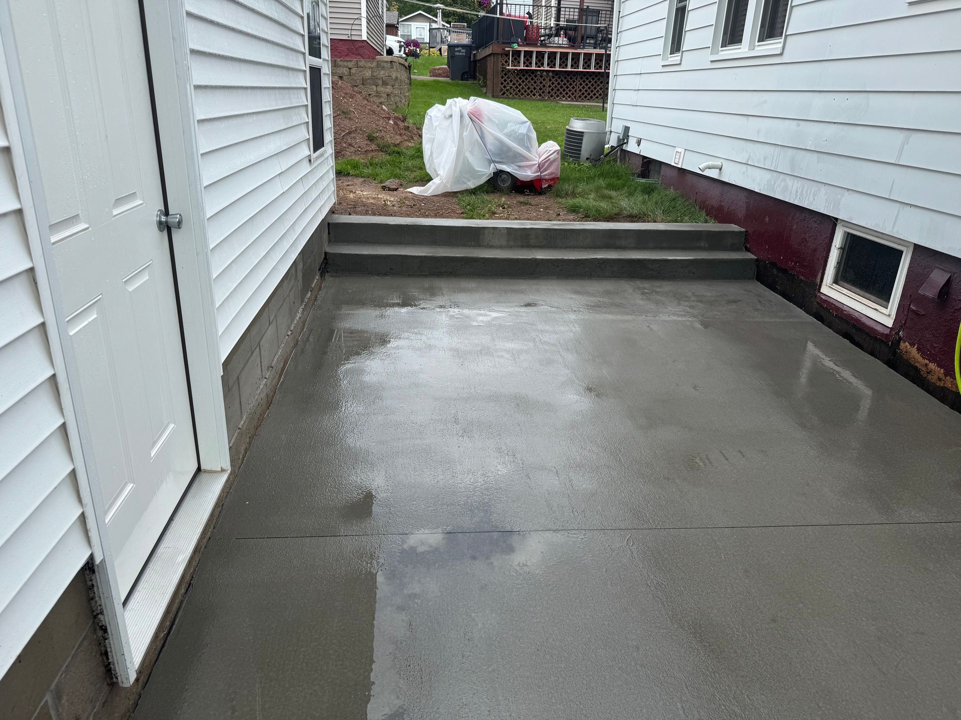 Newly poured concrete patio between two white houses, with steps leading to a yard area.