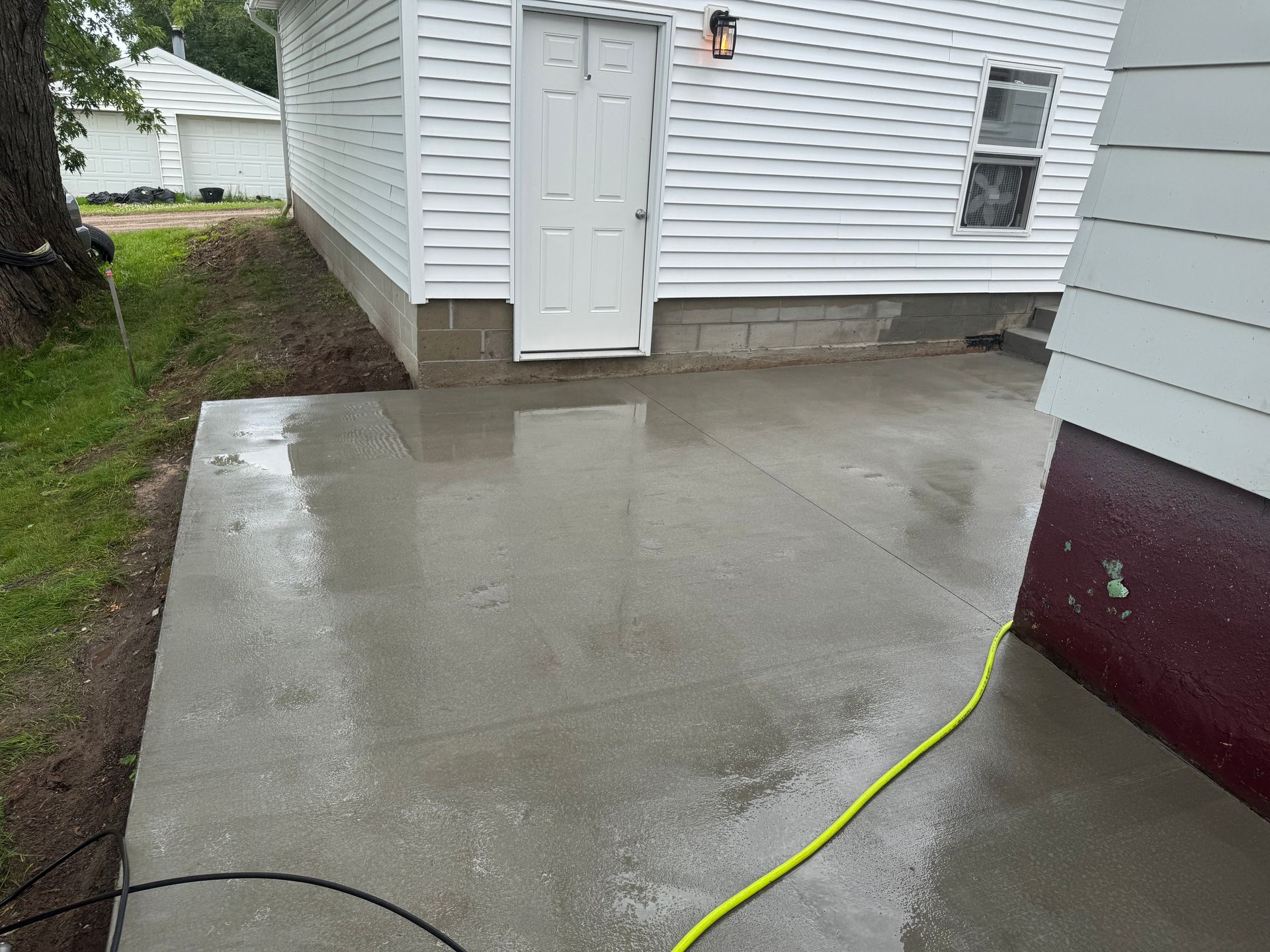 Newly poured concrete patio next to a white house with a door, and a partially visible red wall.