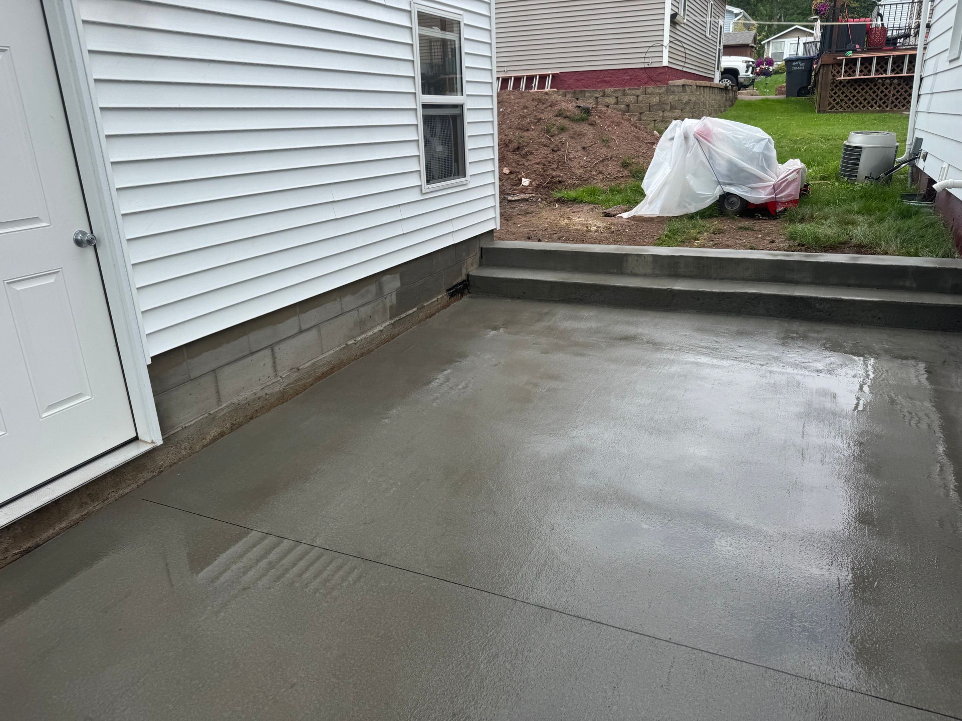 Newly poured concrete patio next to a house with a door, steps, and a small yard.