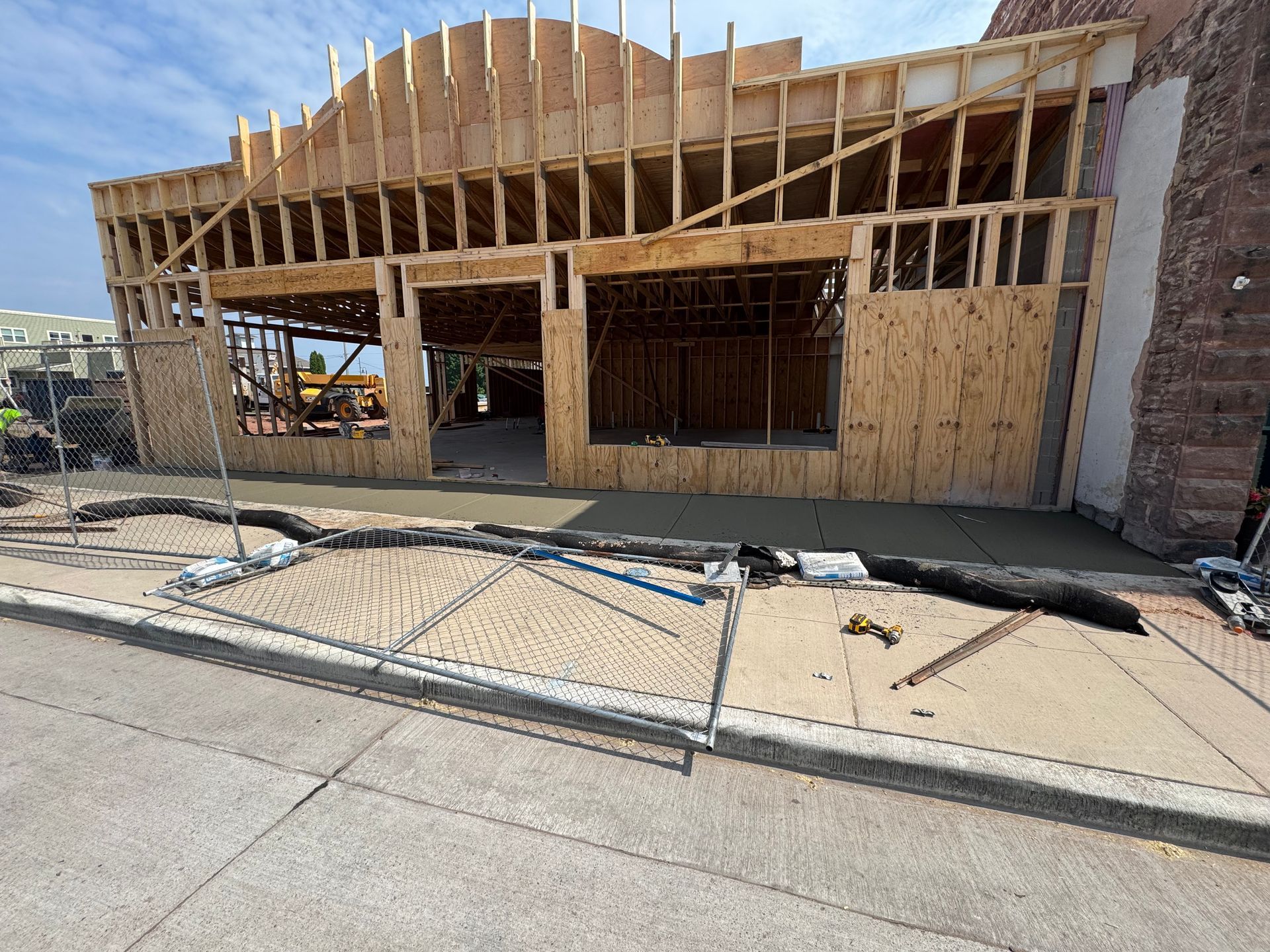 Building under construction with wood framing; exterior view. Concrete sidewalk and street in front.
