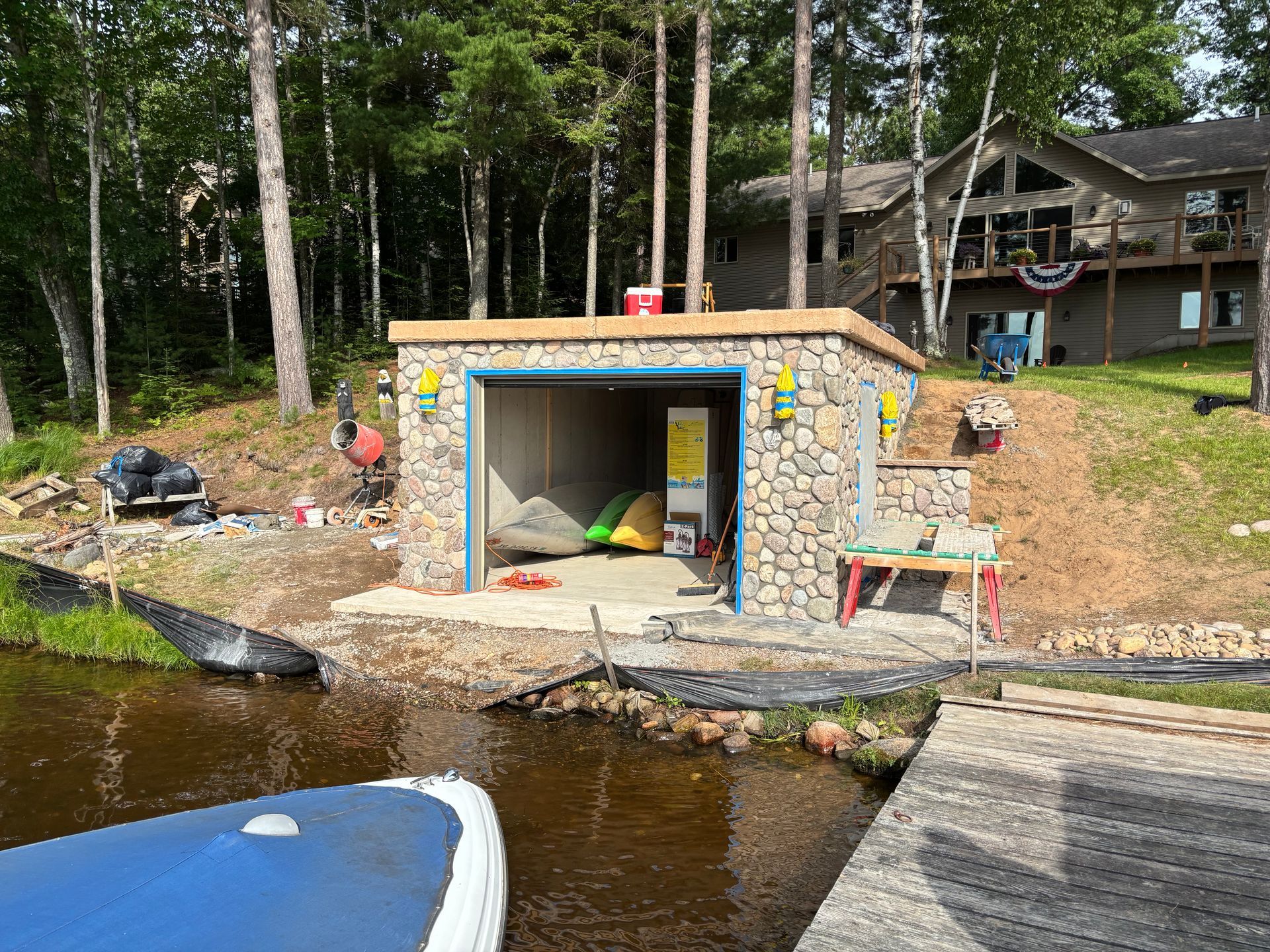 A boathouse with stone exterior on a lake. Kayaks inside, dock to the right, and trees in the background.