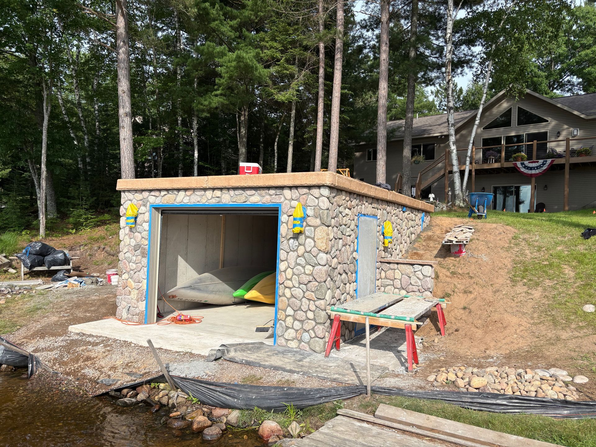 Stone boathouse with kayaks, near a lake and trees. House in background.