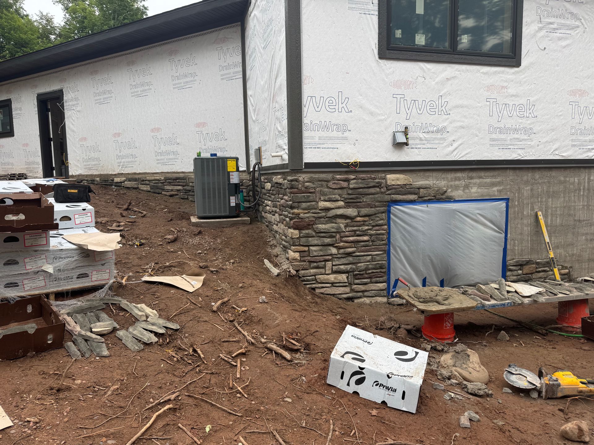 Exterior of a house under construction; exposed wood framing, stone veneer, and dirt with construction materials.