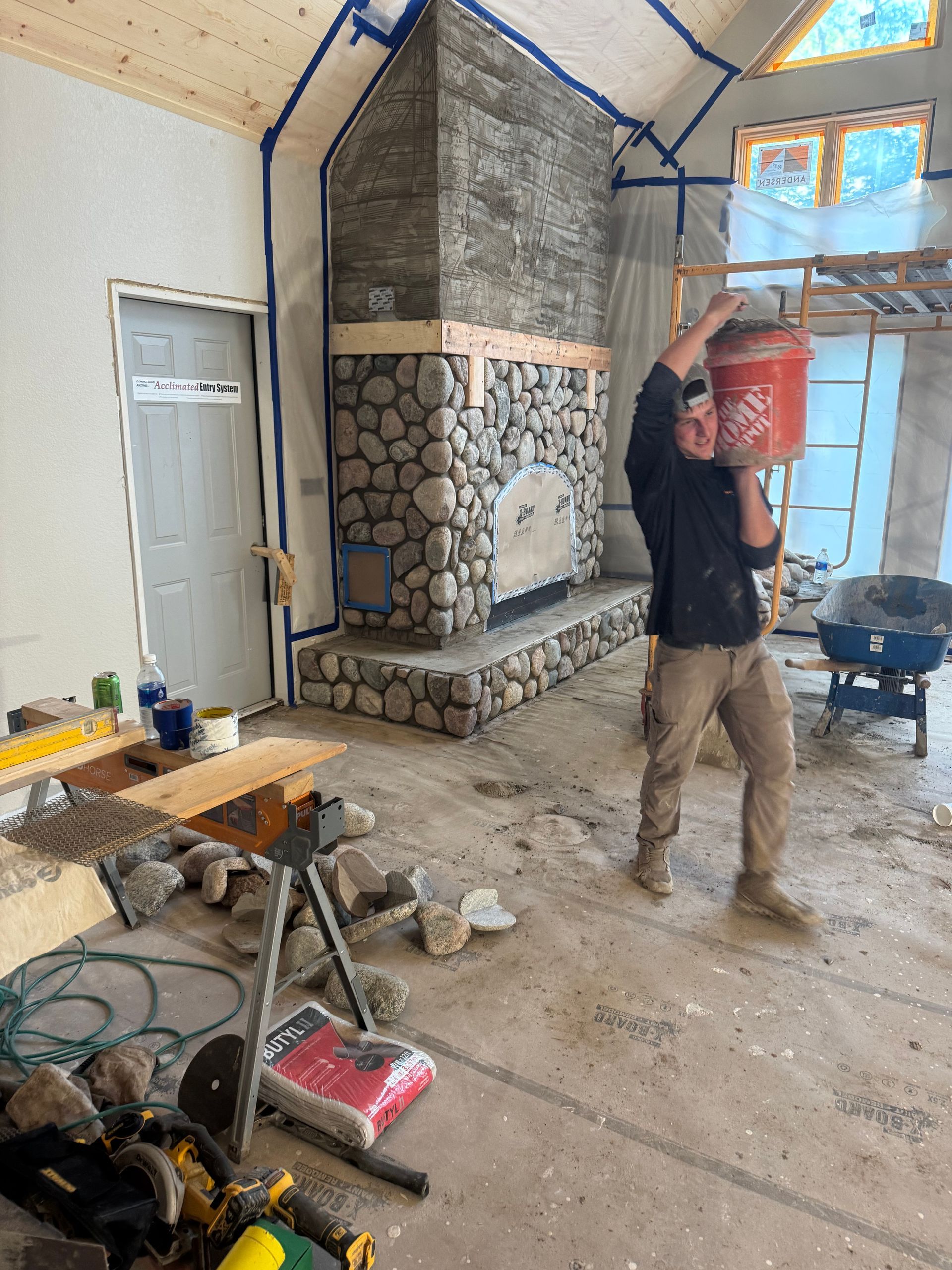 Man carrying a red bucket indoors during construction, near a stone fireplace.