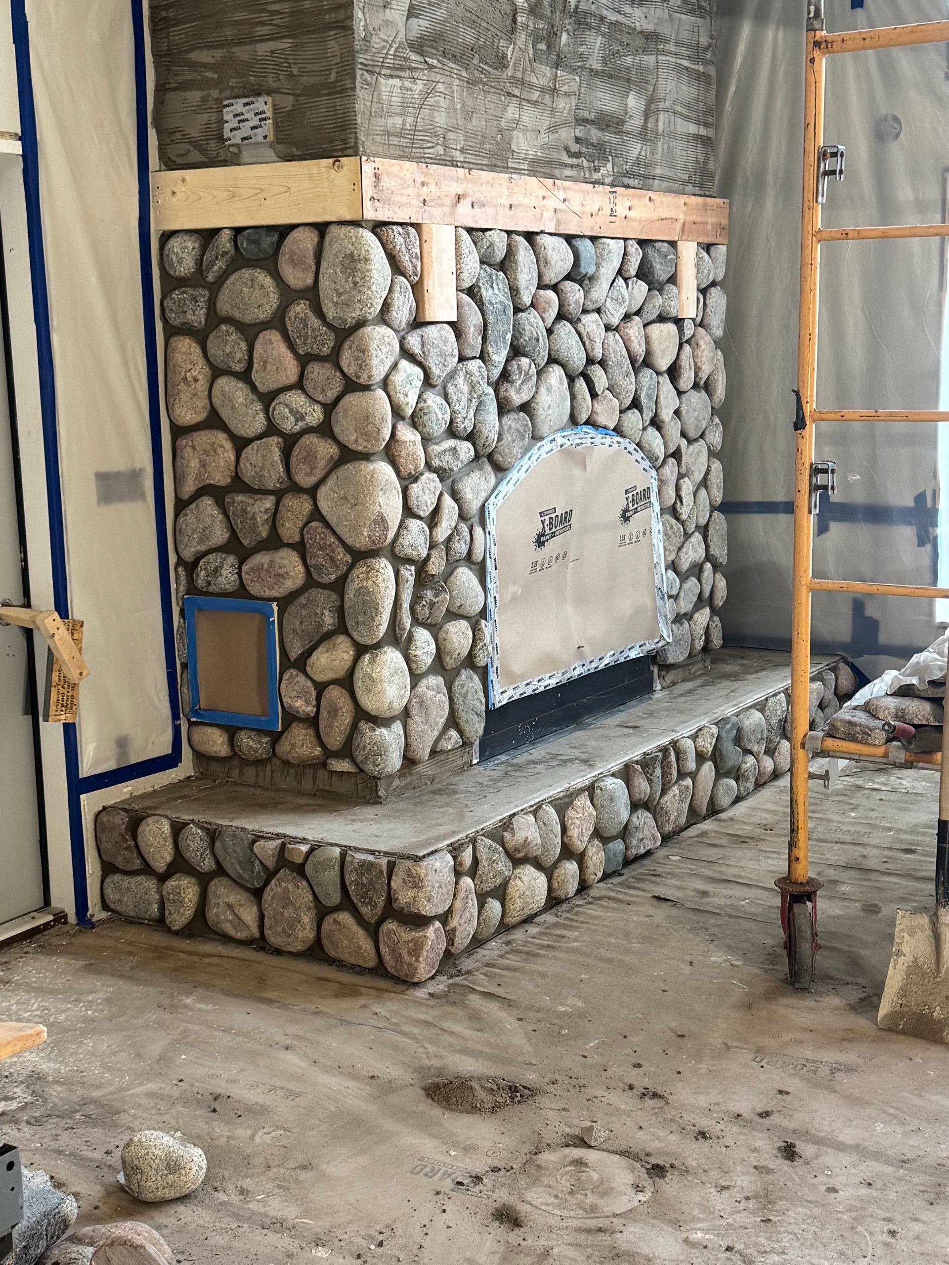 Stone fireplace under construction with a wooden mantle and metal ladder.