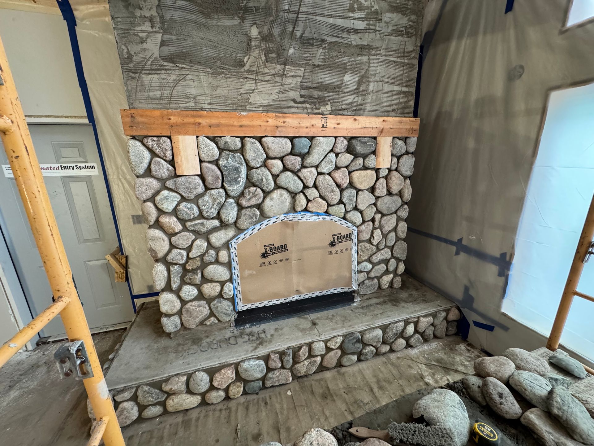 Stone fireplace under construction, with wooden mantle and raw concrete base.