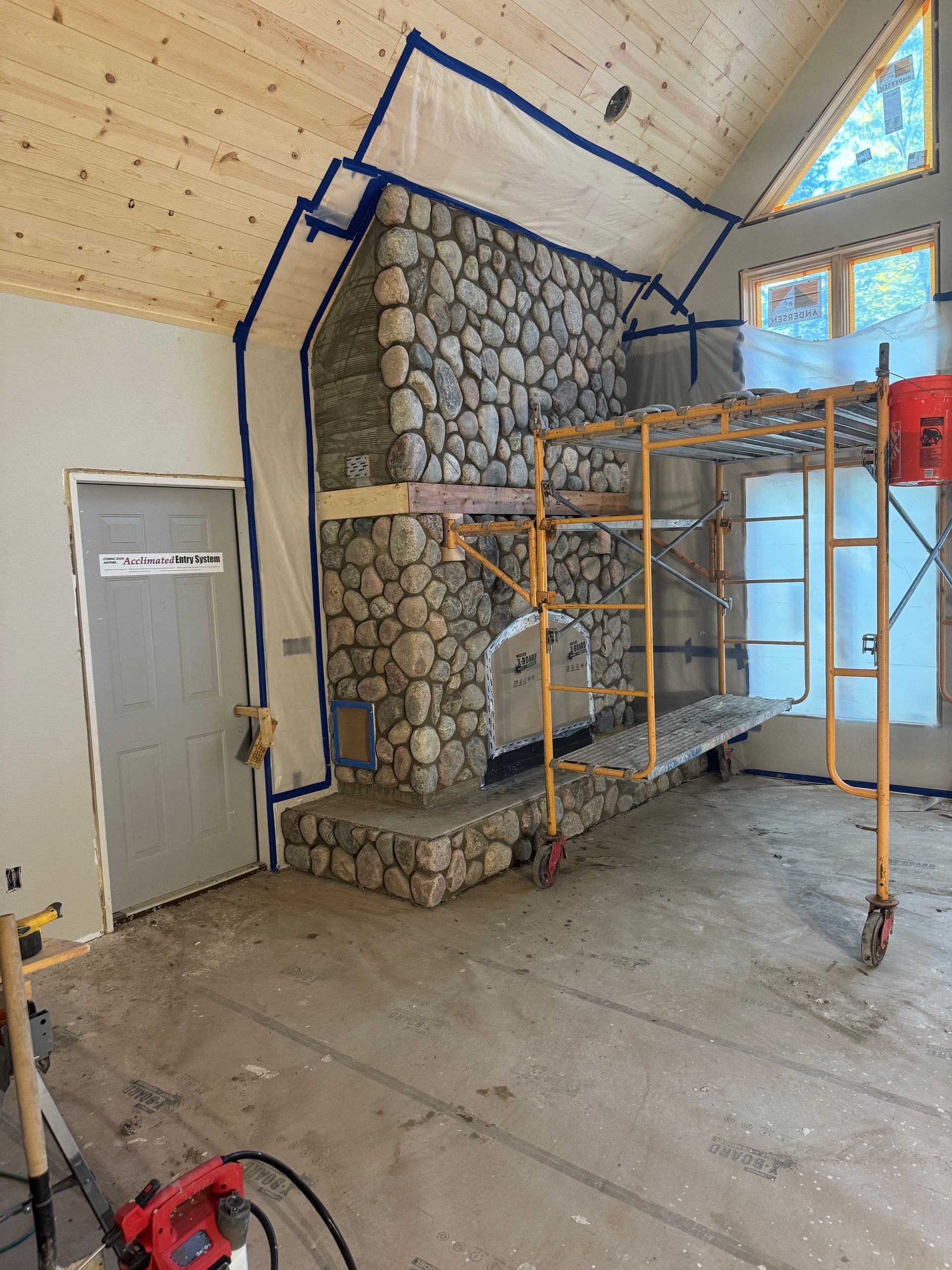 Stone fireplace under construction with scaffolding. Room has gray door and light-colored ceiling.