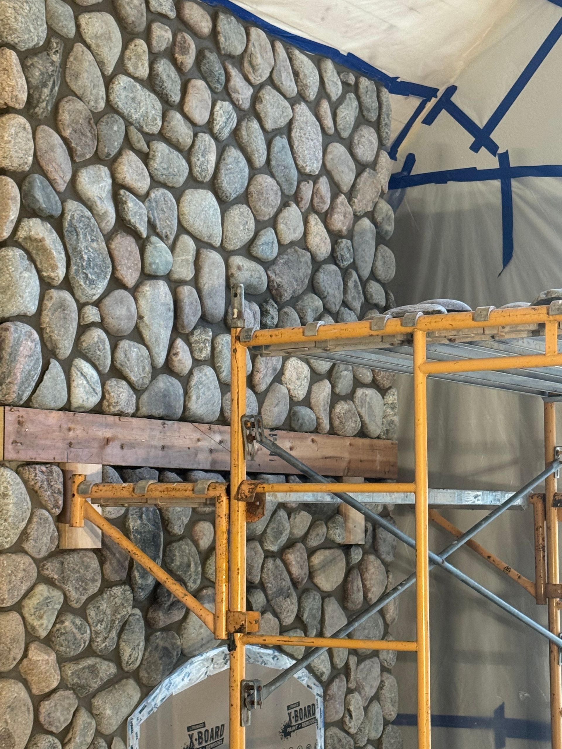 Stone wall construction with a wooden beam and scaffolding. Blue tape is on the ceiling.