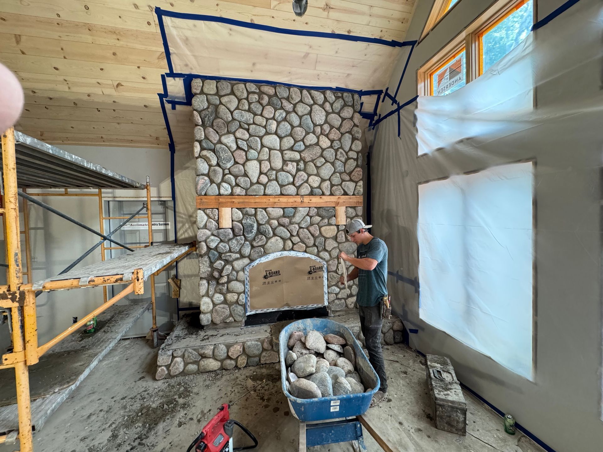 Man building a stone fireplace in a room under construction. Scaffolding, rocks, and tools visible.