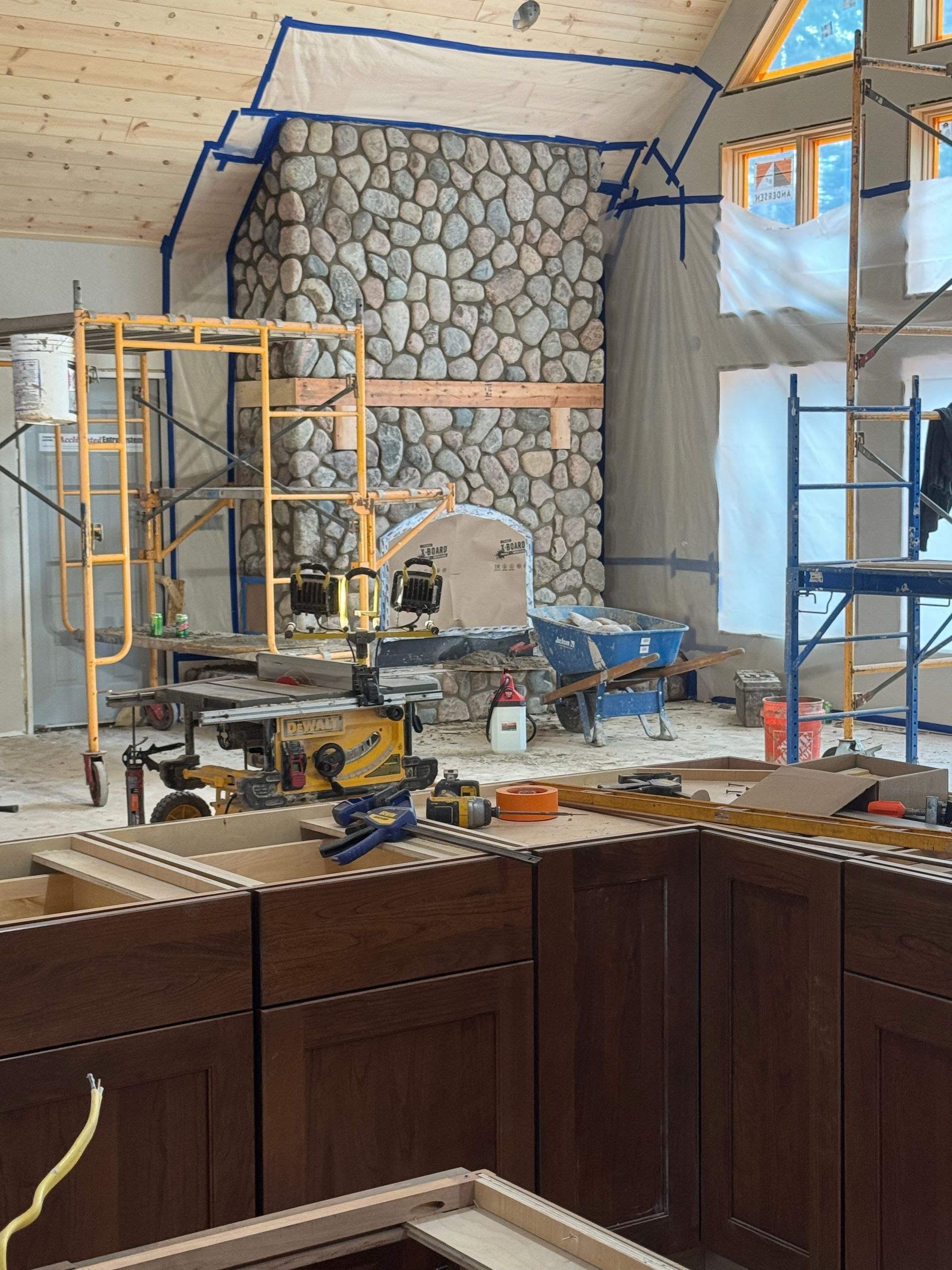 Kitchen renovation in progress, featuring stone fireplace, cabinets, scaffolding, and tools.
