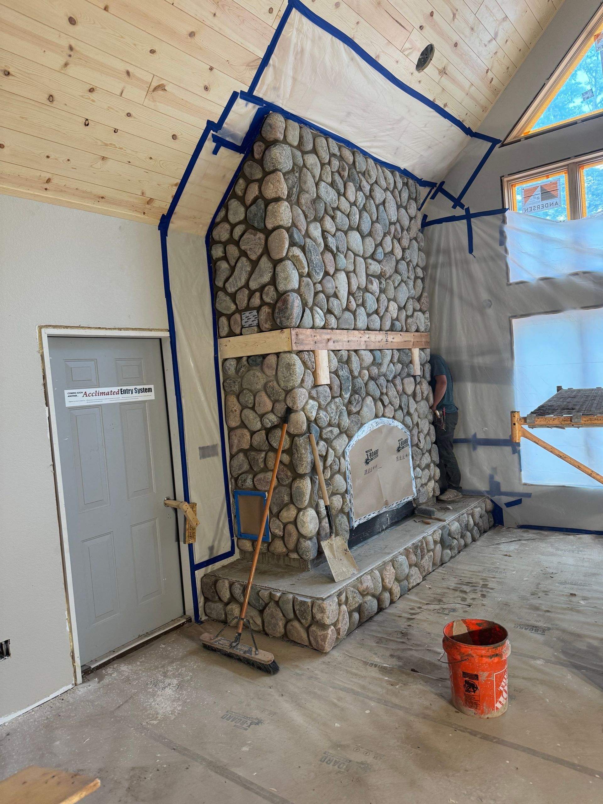 Stone fireplace under construction in a room. Blue tape protects walls, a door is on the left, a broom leans on the hearth, and a bucket is on the floor.