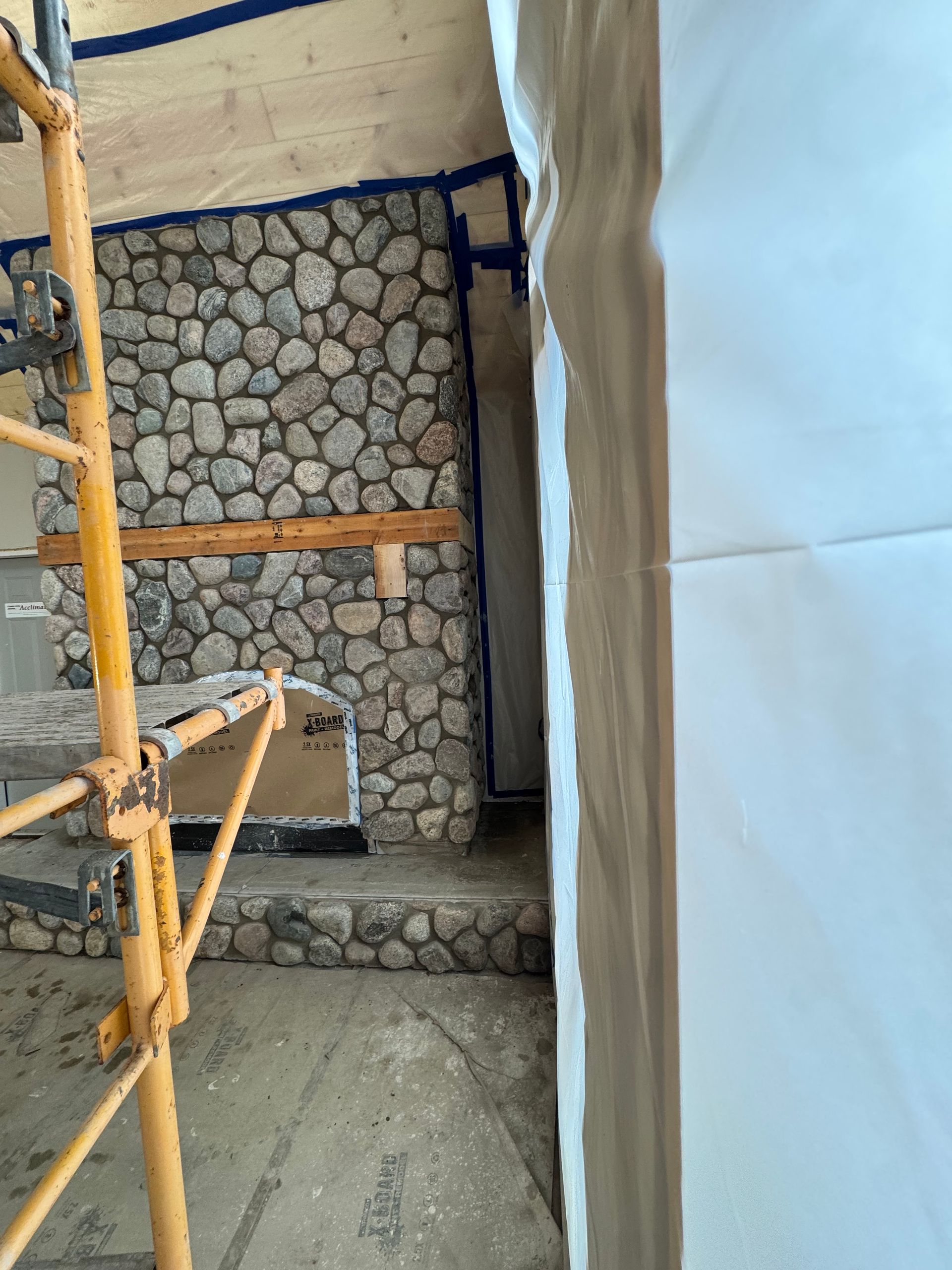 Stone fireplace and steps under construction; scaffolding on the left, partially covered wall on right.