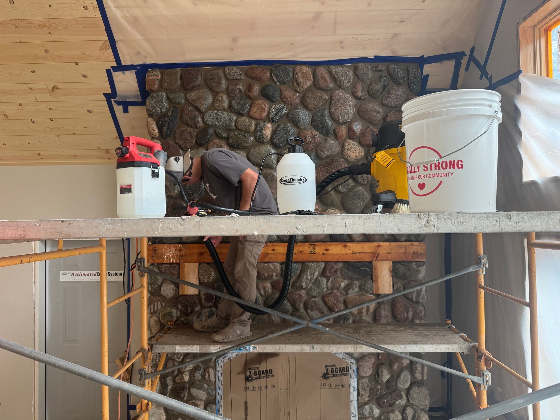 Man working on a stone fireplace using tools, scaffolding. Bucket, vacuum, and paint bottle are nearby.