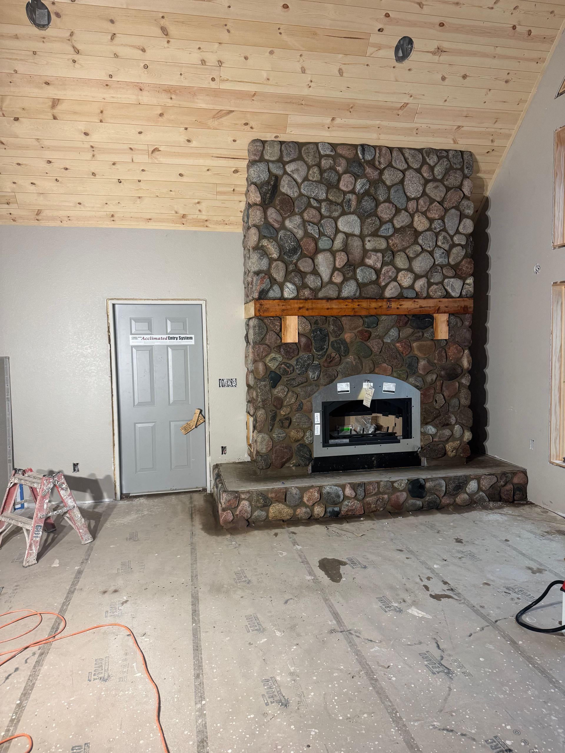 Room under construction with stone fireplace, door, unfinished ceiling and floor.