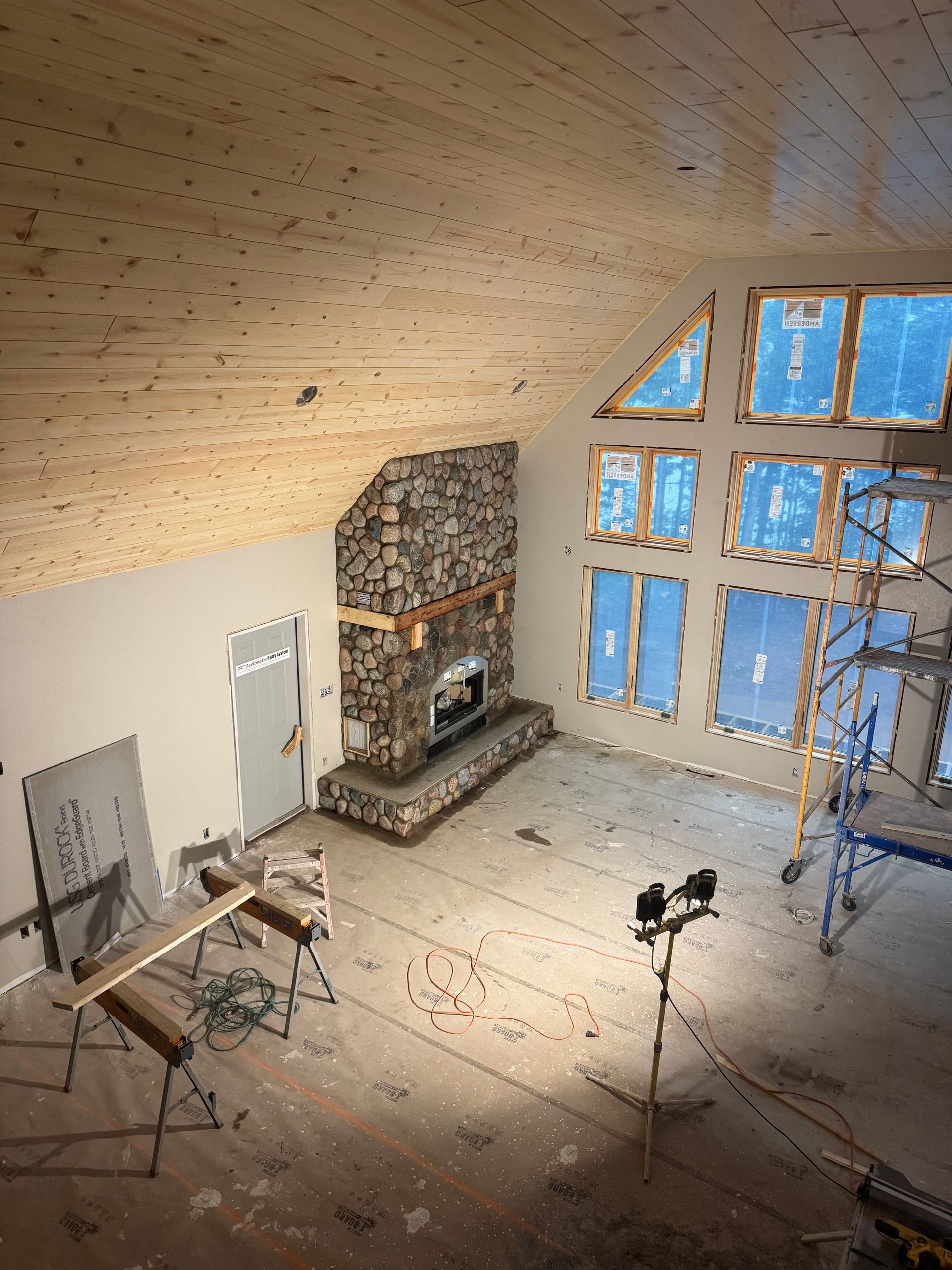 Interior view of a room under construction with a stone fireplace, large windows, and unfinished wood ceiling.