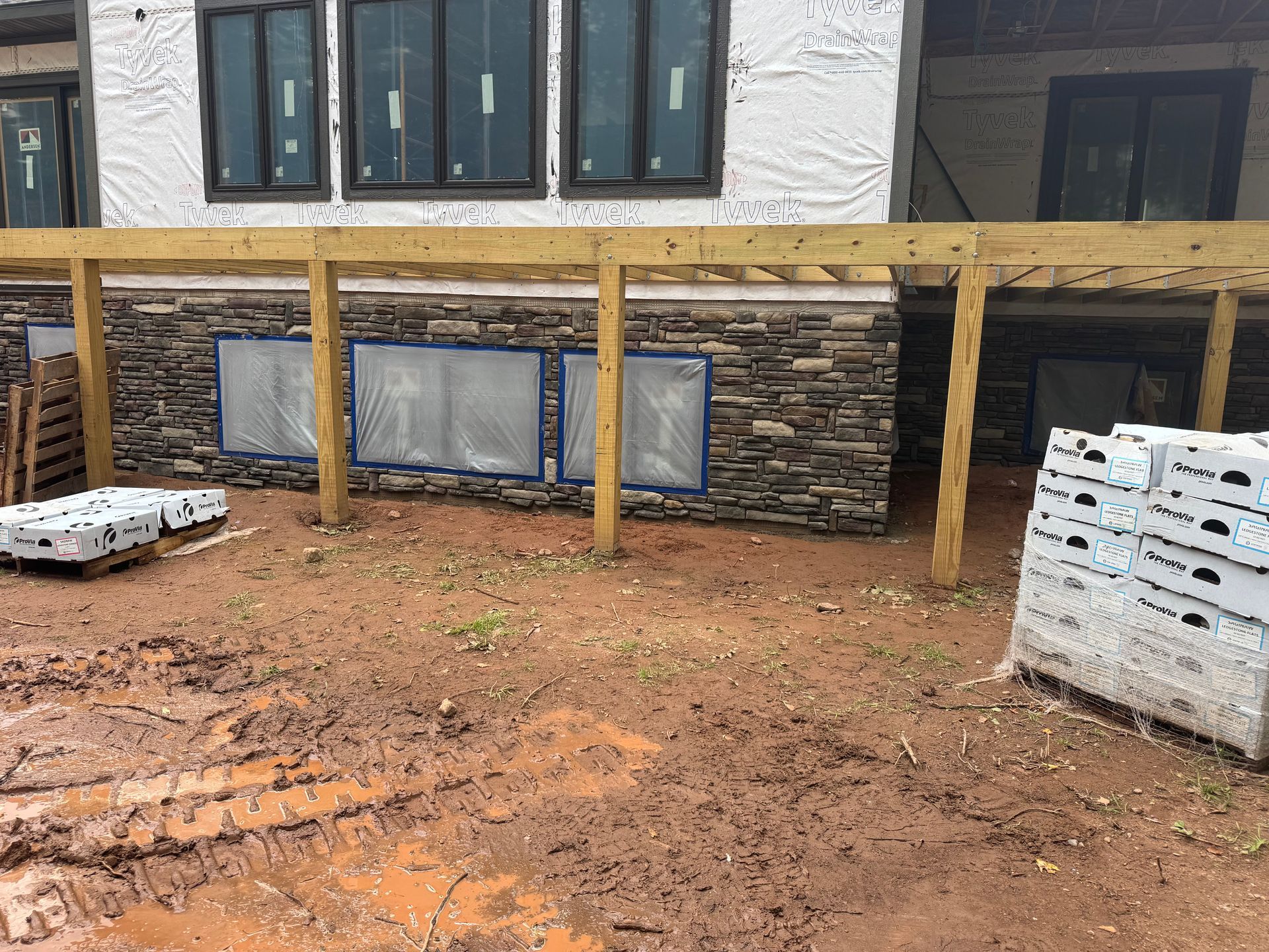 Construction site with stone veneer on a building. Brown wood deck and muddy ground. Windows are covered.