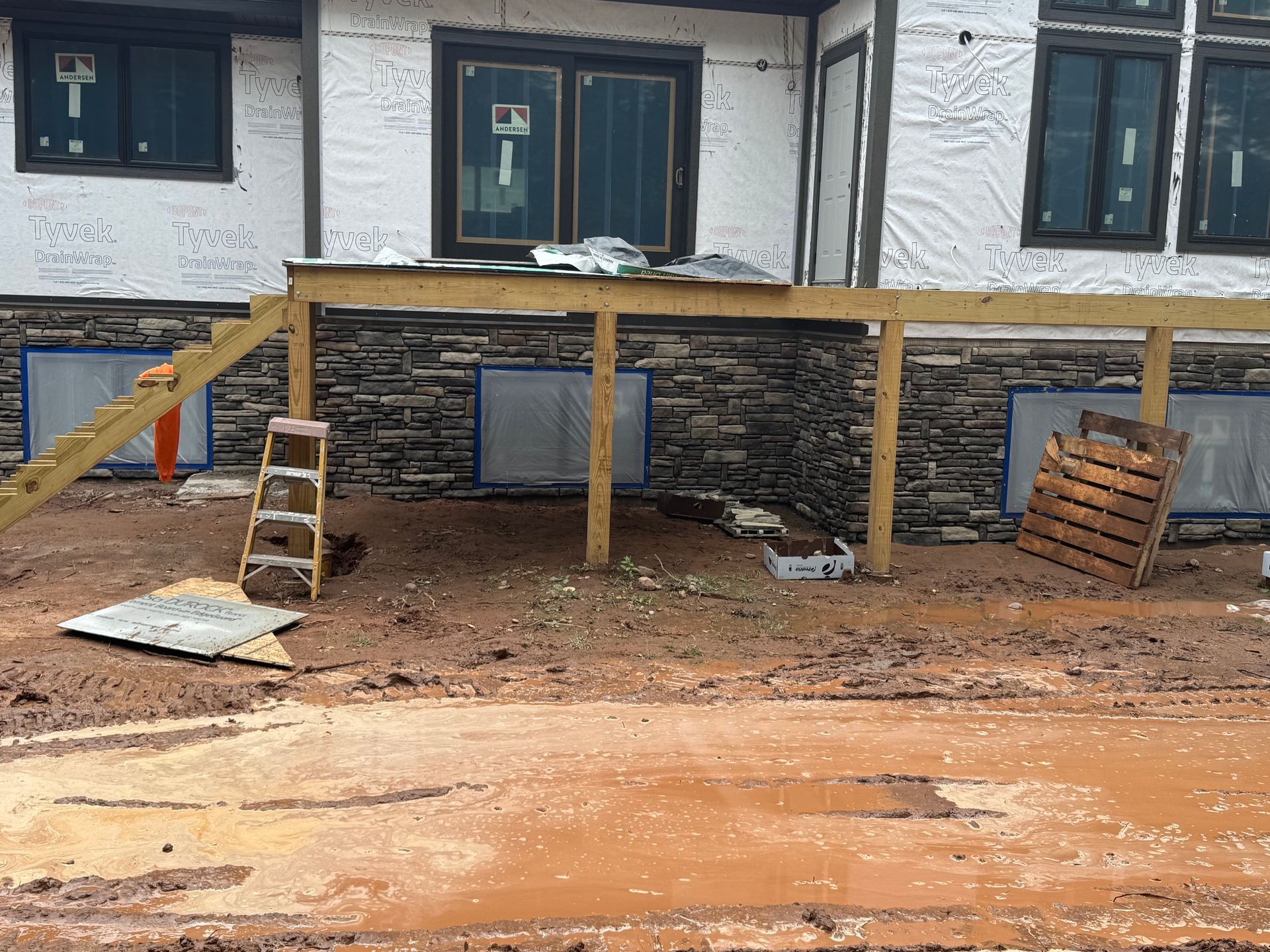 Muddy construction site with a partially built deck, stone veneer, and windows.