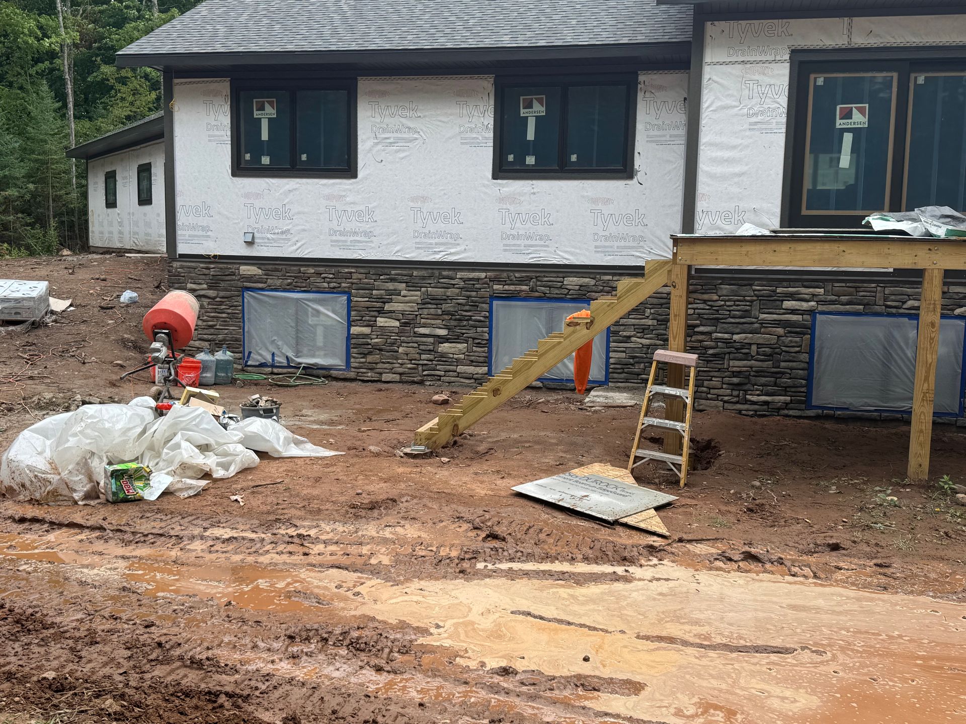 House exterior under construction, mud, stone veneer, windows, deck frame, ladder, and a cement mixer.