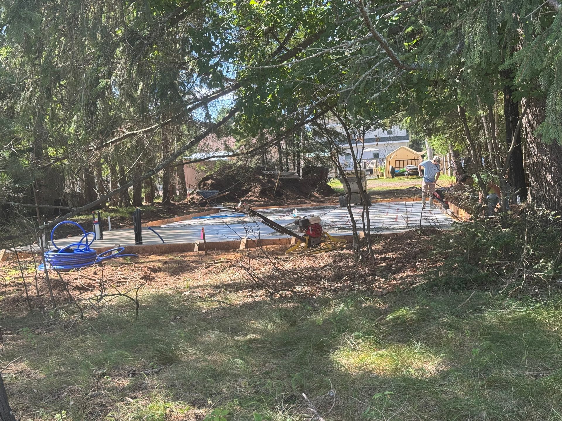 Concrete slab being poured, two people, wooded area, dirt ground, trees, sunny day.