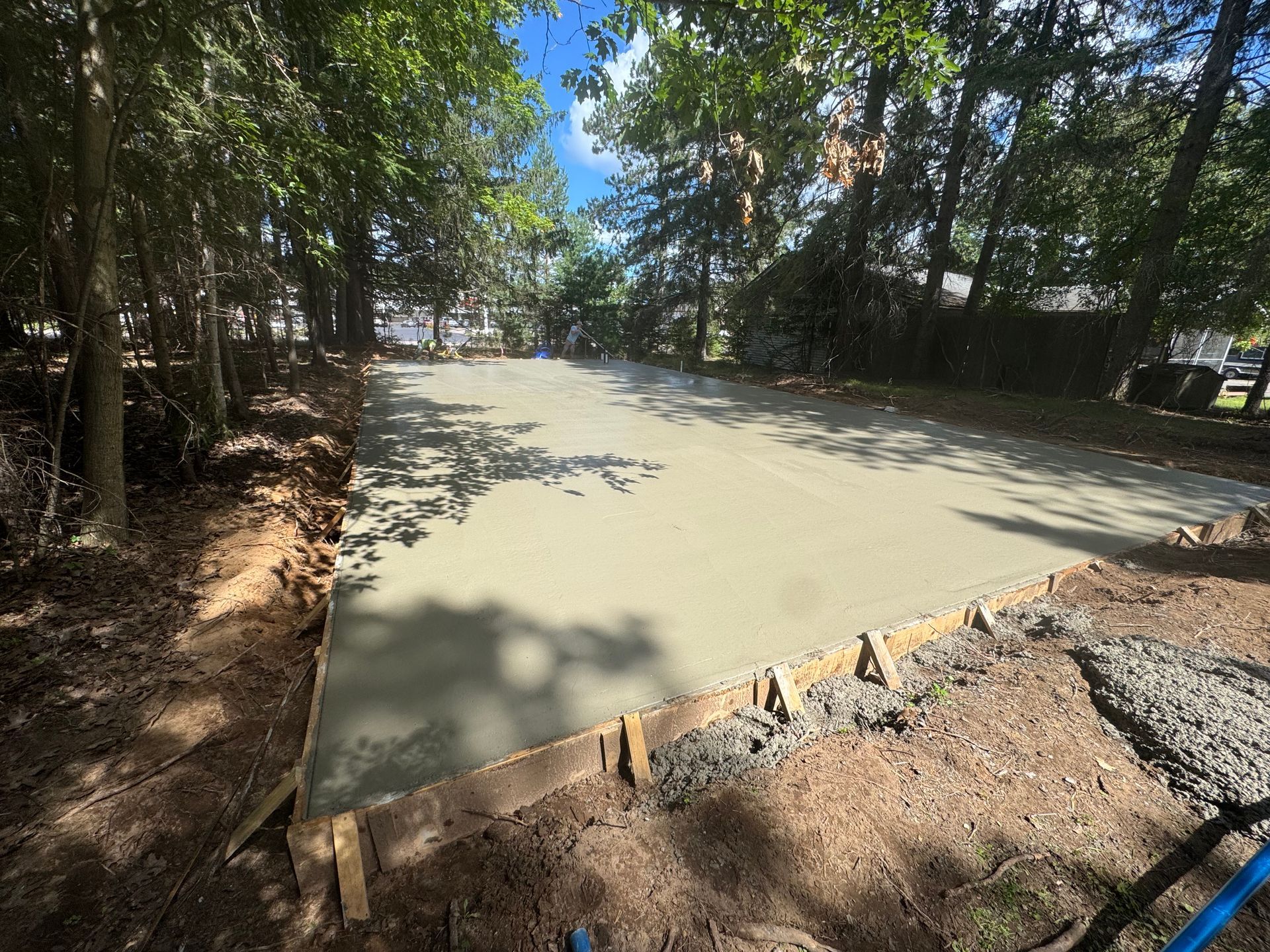 Freshly poured concrete slab bordered by wooden forms and soil, under trees.