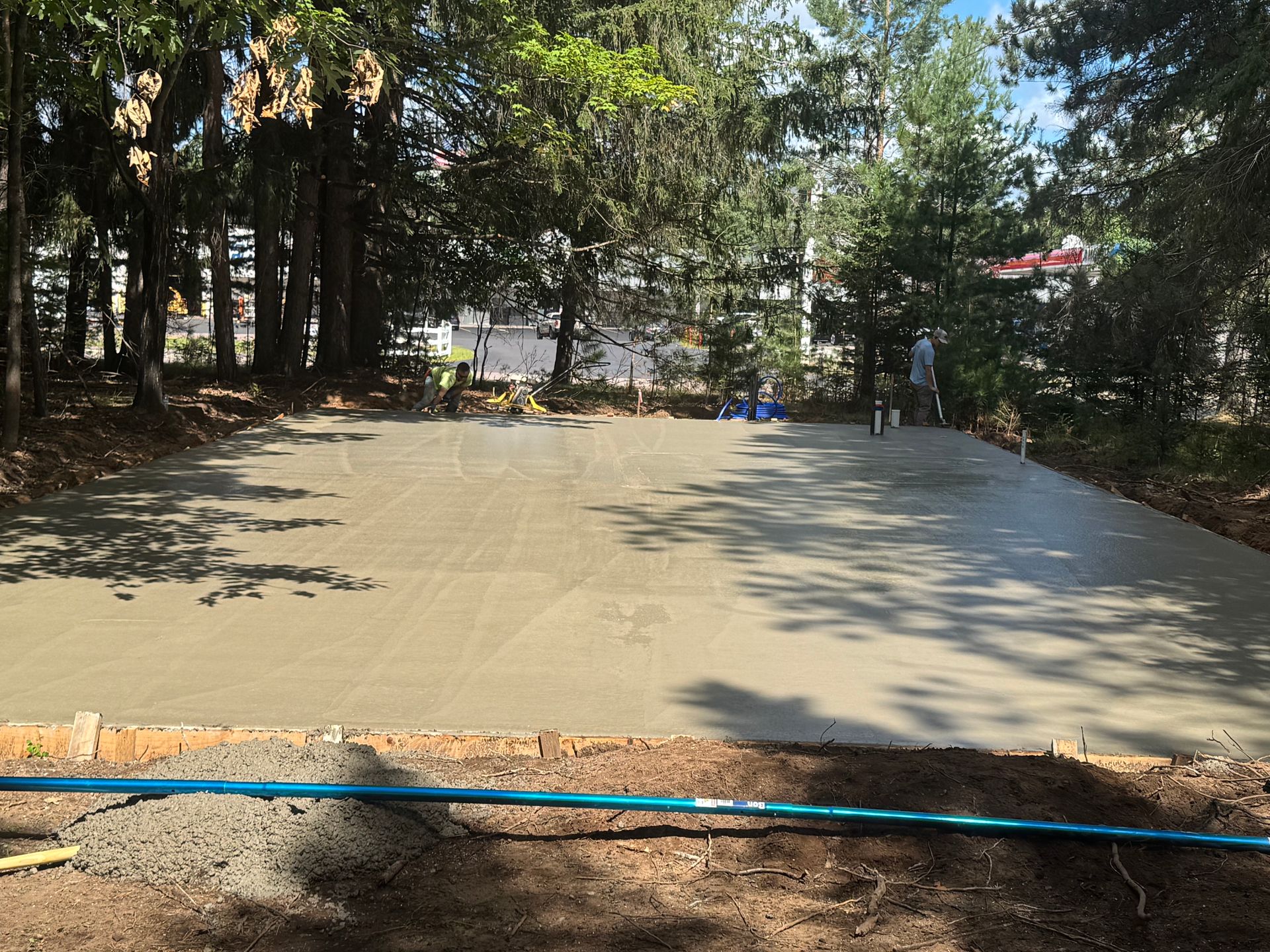 Freshly poured concrete slab in an outdoor setting, surrounded by trees and construction workers in the distance.