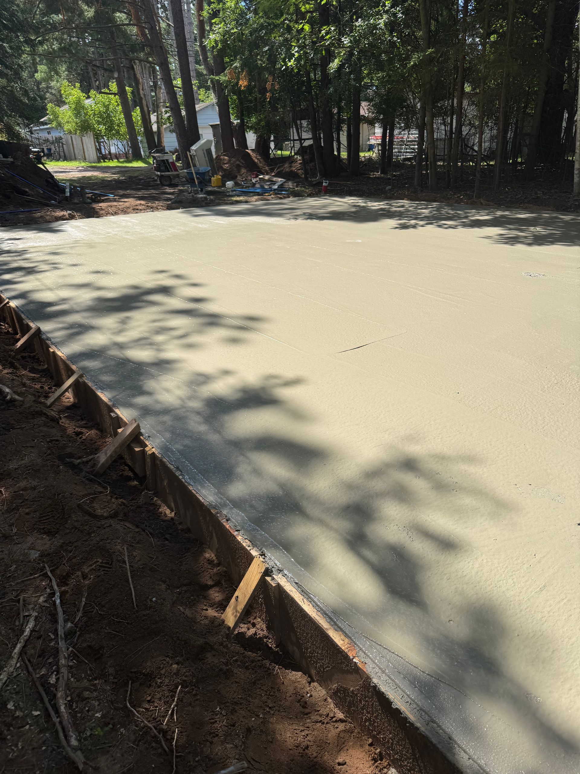 Freshly poured concrete slab with wood formwork on the edge, shaded by trees in the background.