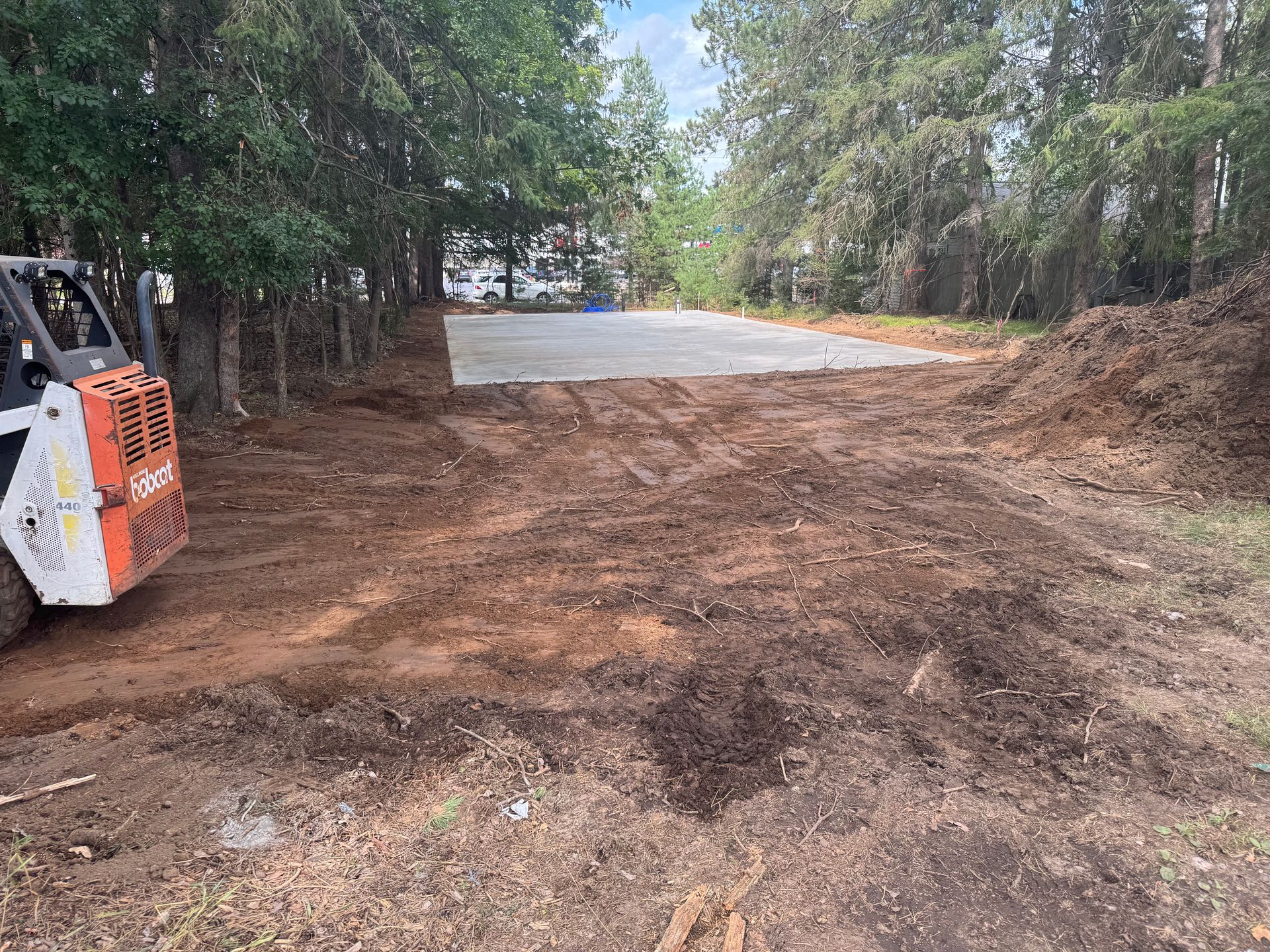 A small excavator on muddy ground, clearing an area near trees and a gravel pad.