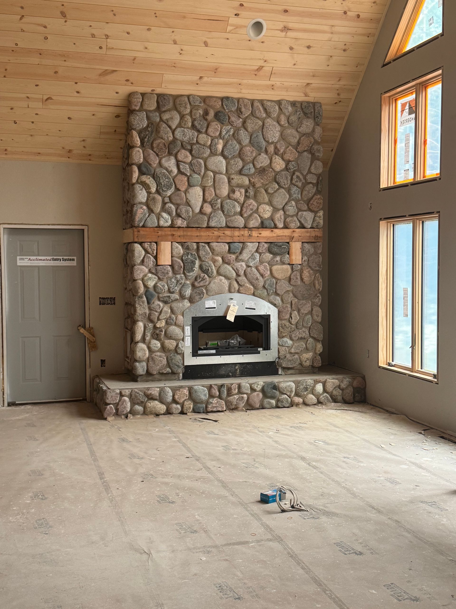 Stone fireplace in a room under construction with windows and a wooden ceiling.