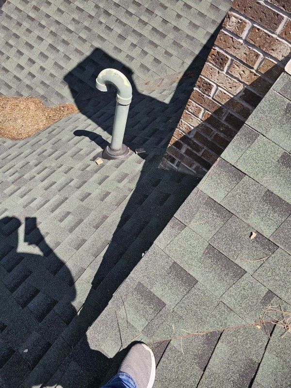 Person's shadow and foot on a green shingled roof, near a vent pipe and brick chimney, sunny day.