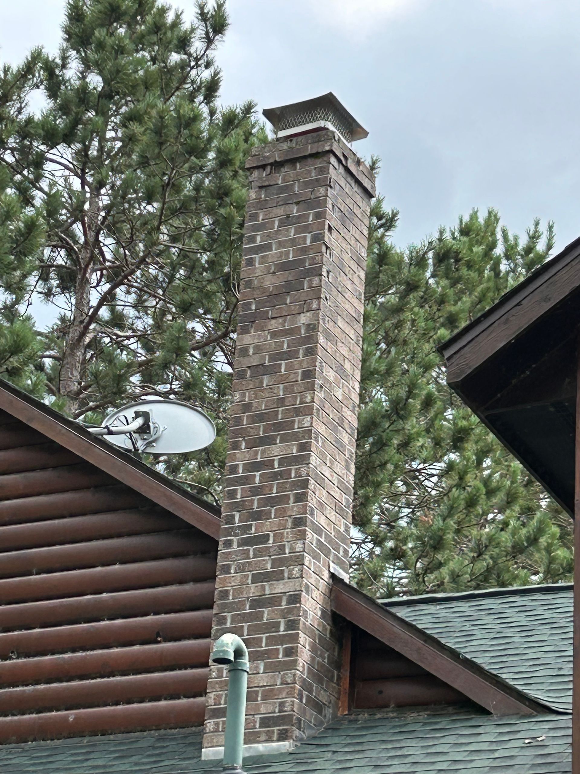 Brick chimney with cap extending from the roof of a log cabin, satellite dish visible.