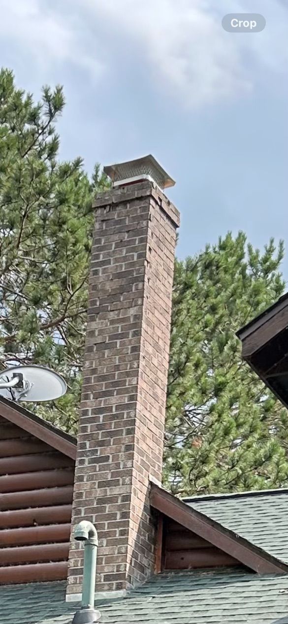 Brick chimney with a cap, on a rooftop, surrounded by trees and a cloudy sky.