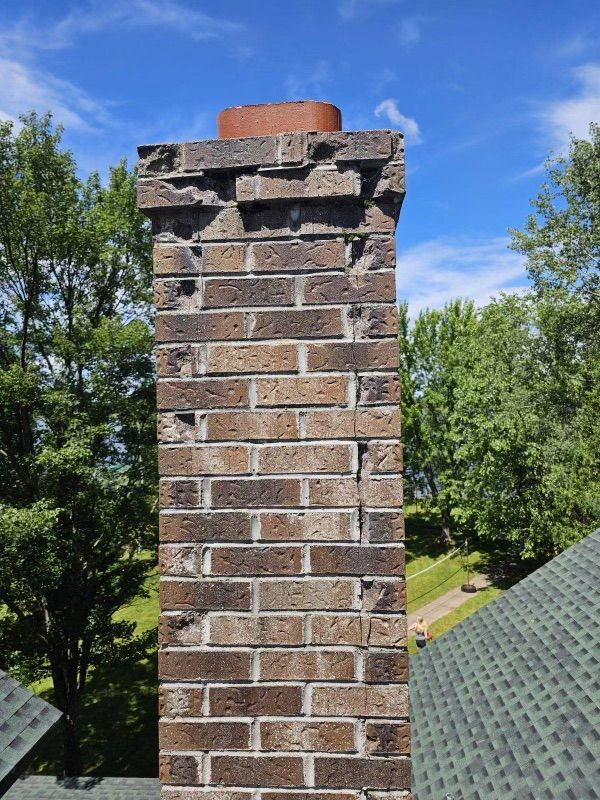 Brick chimney, weathered and damaged, against a blue sky with trees and green rooftop visible.