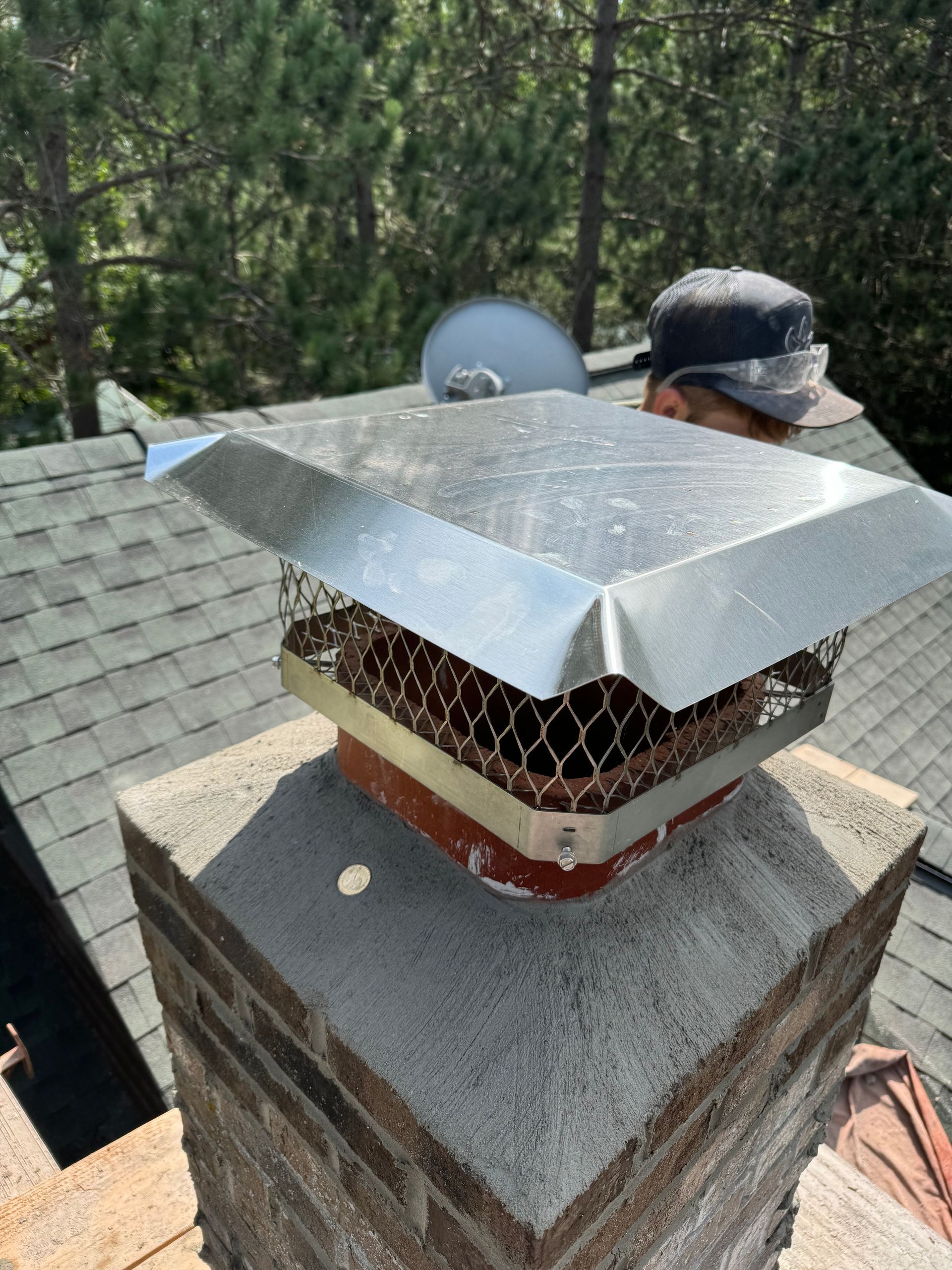 Person installing a metal chimney cap on a brick chimney on a roof.