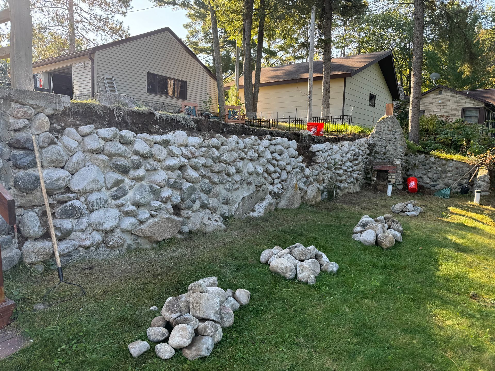 A stone retaining wall being constructed in a grassy yard, with houses and trees in the background.