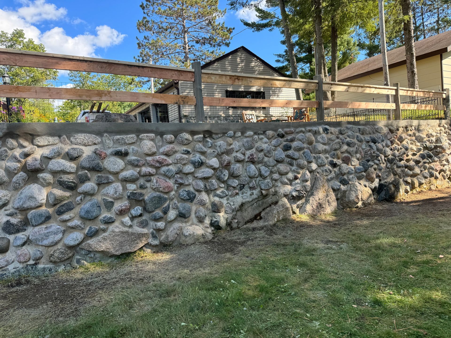 Stone retaining wall with a wooden fence. Building in the background. Green grass in the foreground.