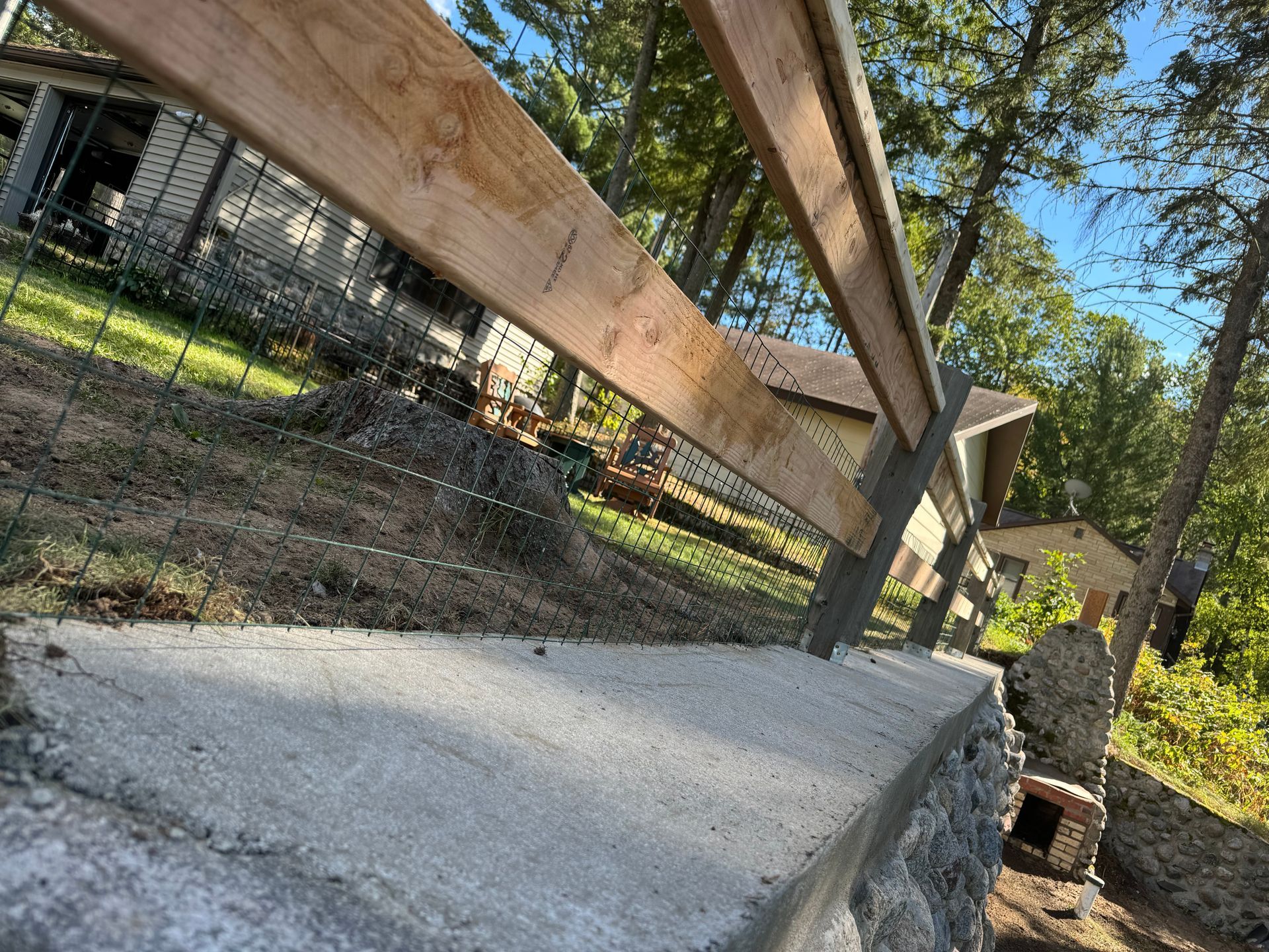 Wooden fence atop a concrete and stone retaining wall, with a yard and buildings in the background.